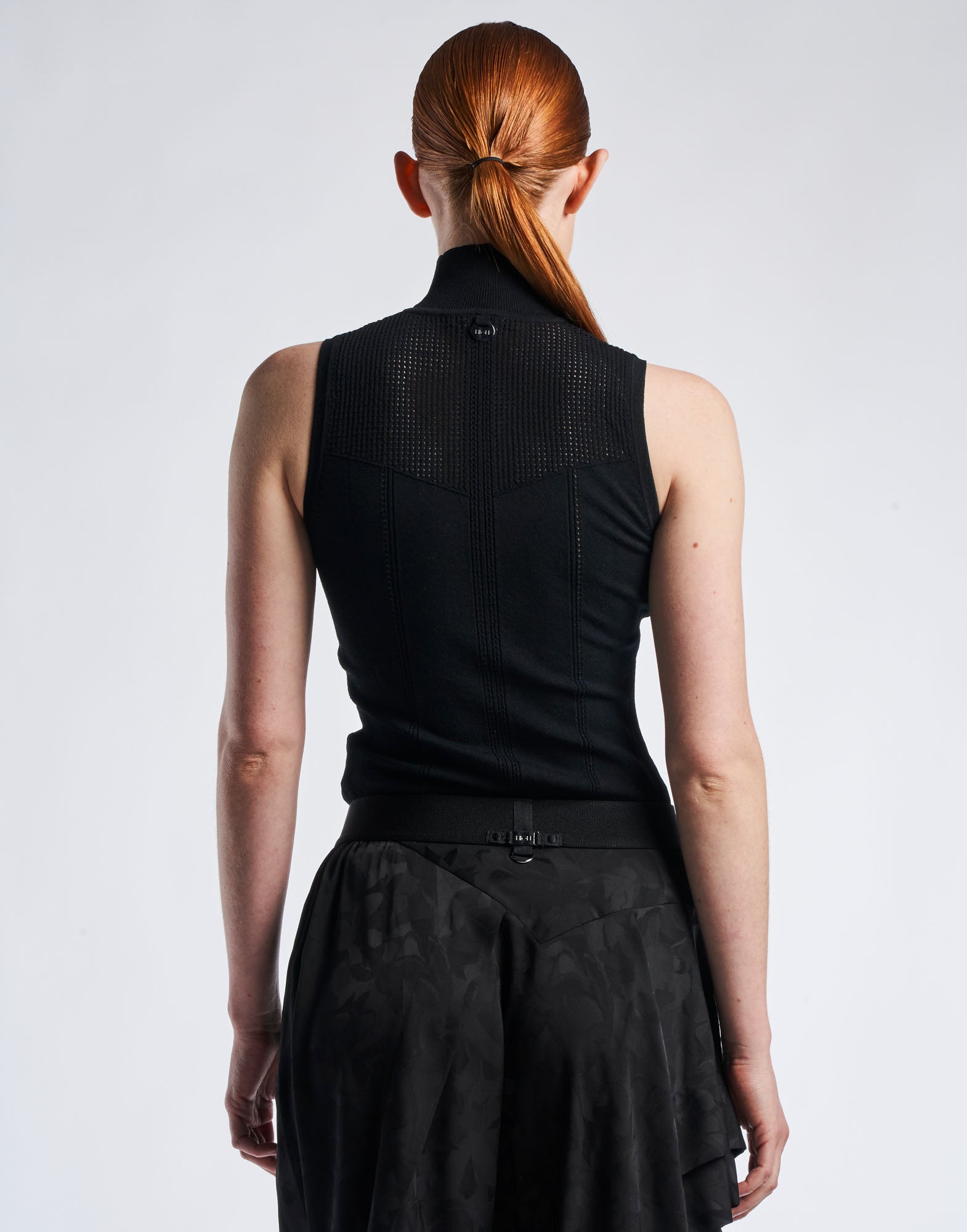 Back view of a woman wearing the sleeveless black GUISE top with high collar, perforated yoke, and vertical details. Soft technical knit.