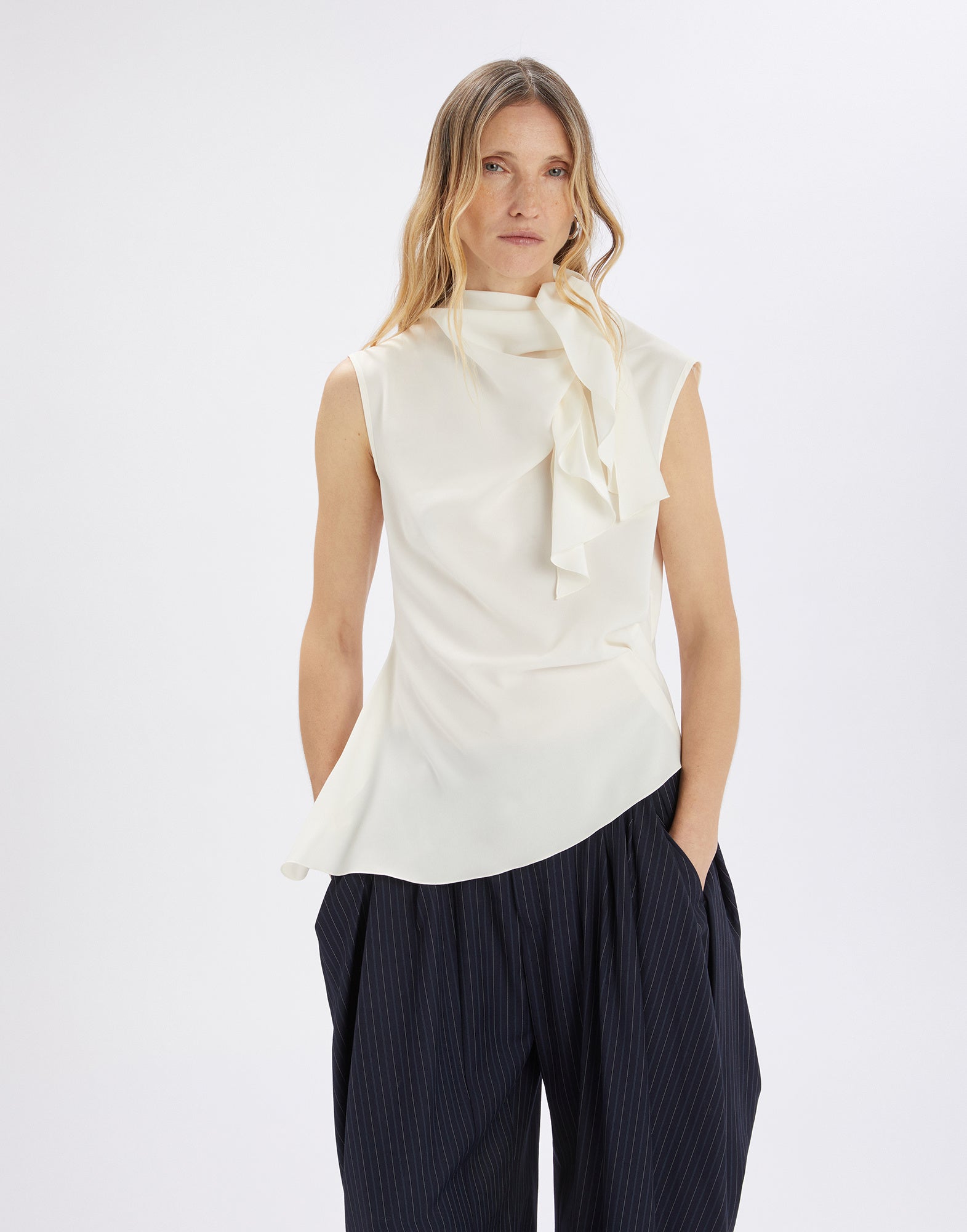 Front view of the WALTON: A woman wears a sleeveless ivory satin top with a side jabot collar and an elegantly draped asymmetric hem.