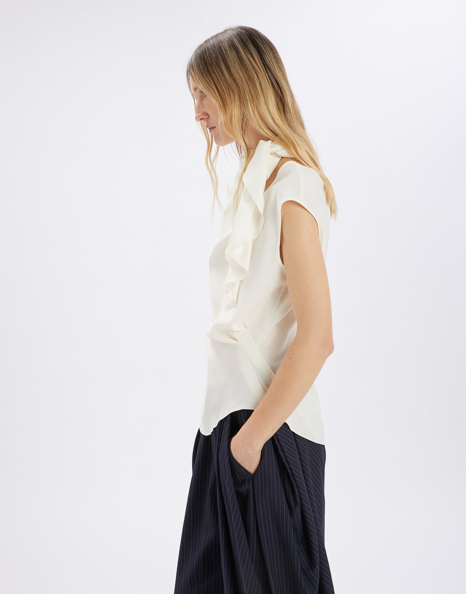 Side view: Woman wears WALTON sleeveless top made of flowing, cream satin with asymmetric hem and side jabot collar. Light and elegant.