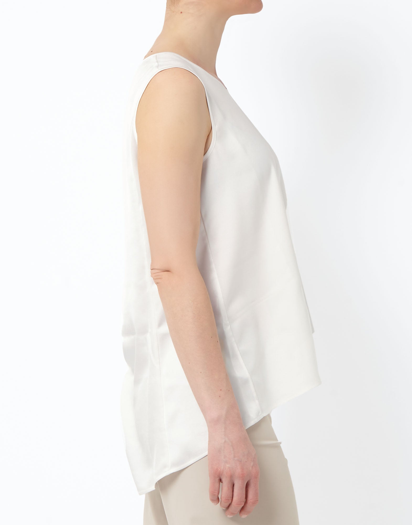 Seitenansicht: Modell trägt das ärmellose Top CUE aus leicht glänzendem, technischem Satin in Off-White, mit rundem Ausschnitt und längerem, weich fallendem Rücken, kombiniert mit hellen Hosen.