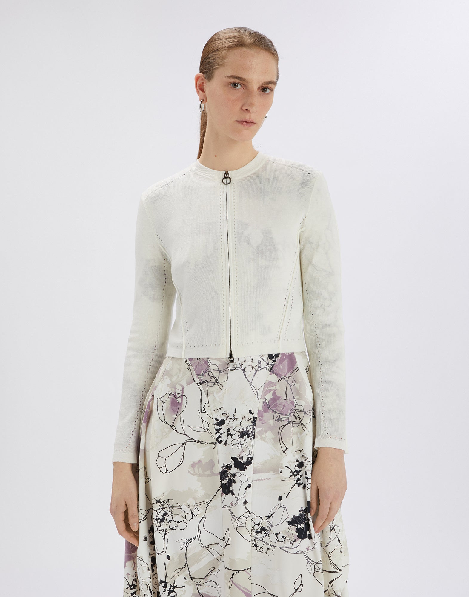 Front view: A woman wears the SKITTER Cardigan in off-white with openwork pattern, crewneck, metal zip closure, and long sleeves, paired with a printed dress.