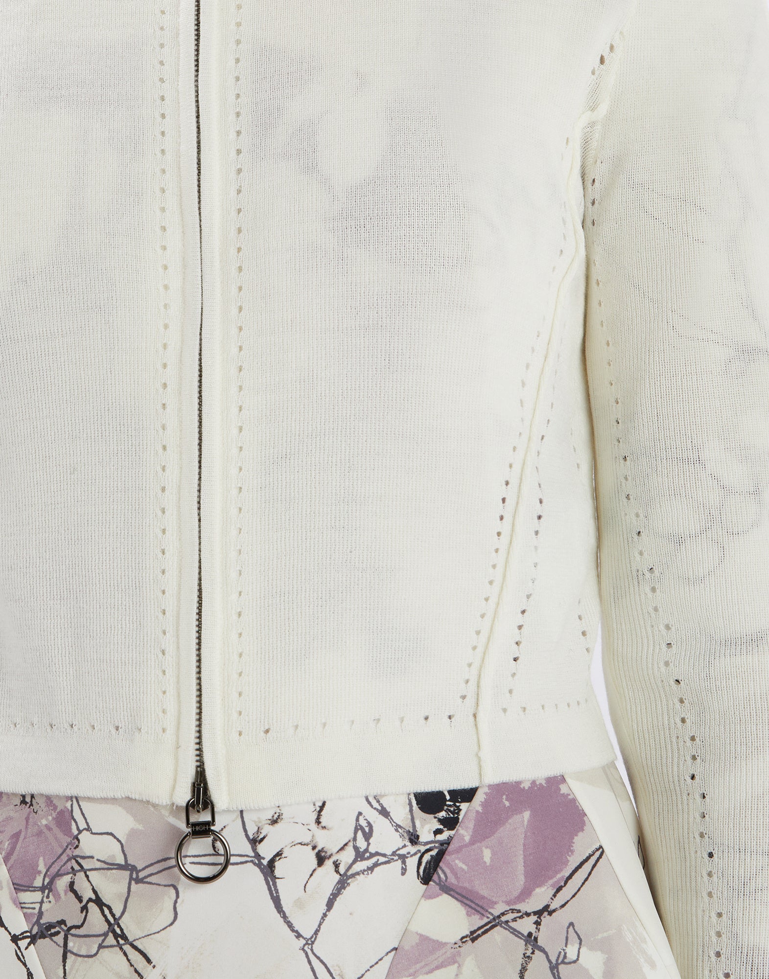 Detailed view of SKITTER shows the front section of a white cardigan with fine openwork pattern, metal zipper, and ribbed cuffs over a patterned skirt.