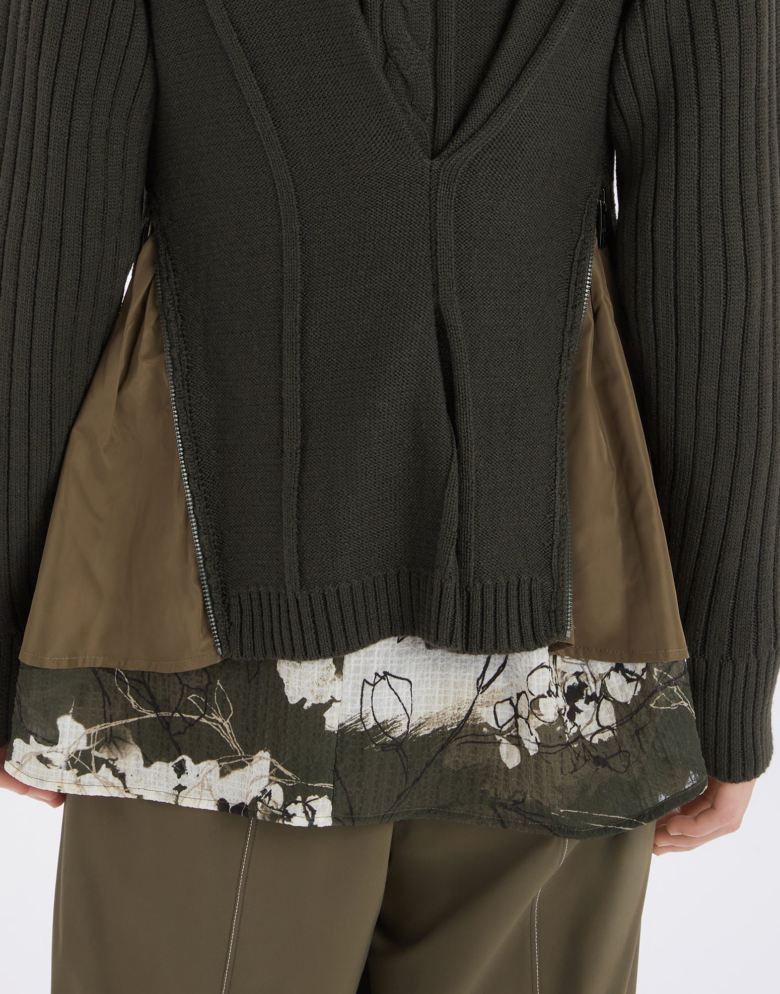 Detailansicht der FREIZE Strickjacke von hinten, zeigt olivgrünes Rippen- und Zopfmuster mit seitlichen Reißverschlüssen und Nylon-Einsätzen über floraler Bluse.