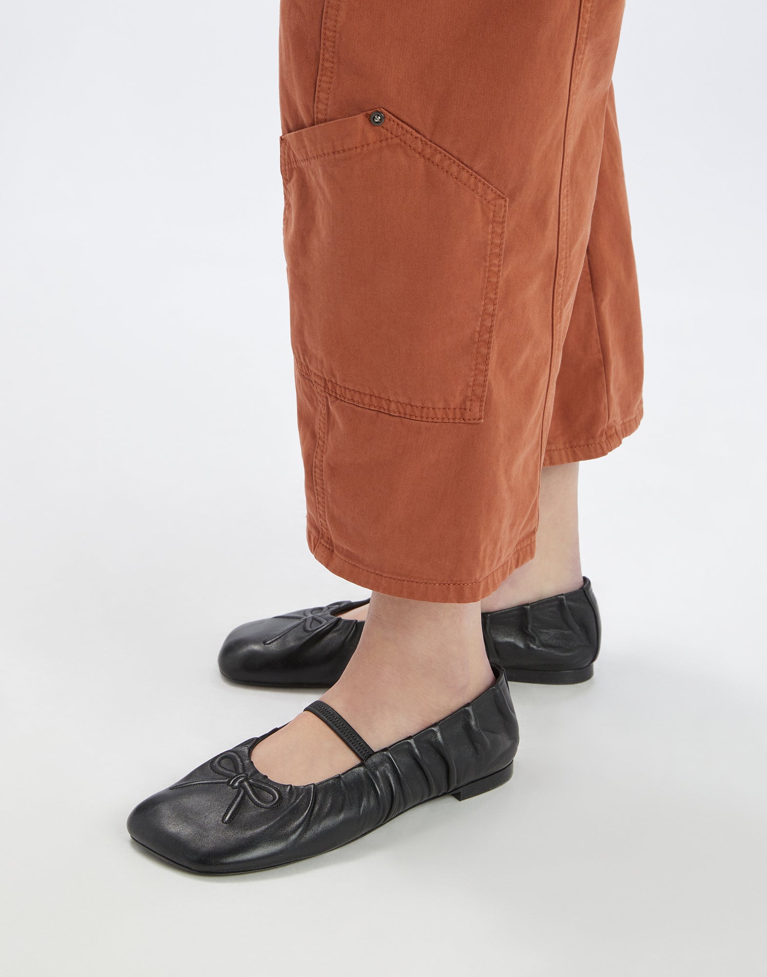 Detailansicht; Person trägt FURORE Hose in rostbrauner Farbe mit aufgesetzter, eckiger Tasche am unteren Bein, sichtbare Nähte, kombiniert mit schwarzen Ballerinas.