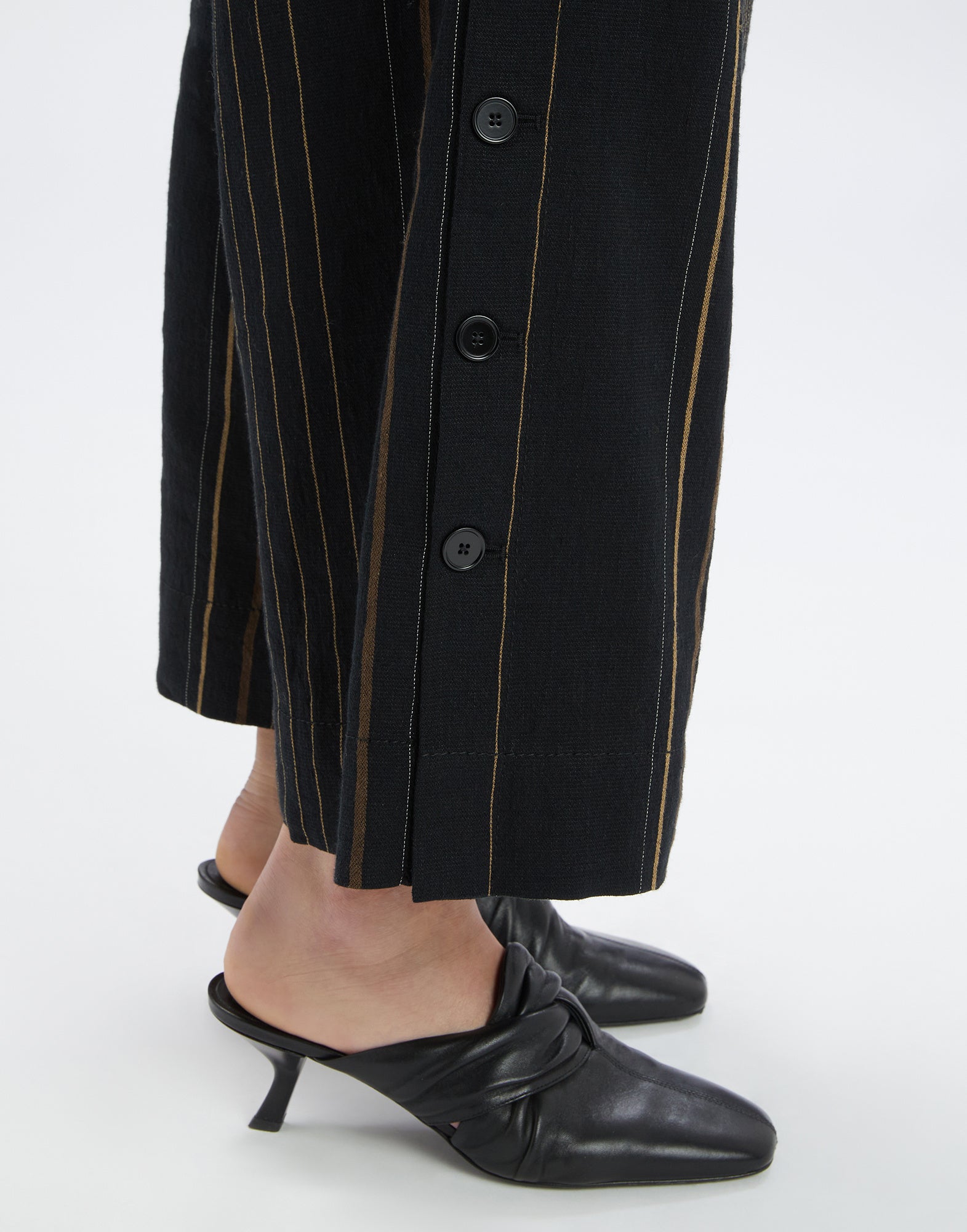 Detailansicht der SWALLOW Hose zeigt den Beinabschluss mit seitlicher Knopfleiste, vertikalen Streifen auf schwarzem Hintergrund und schwarzem Absatzschuh.