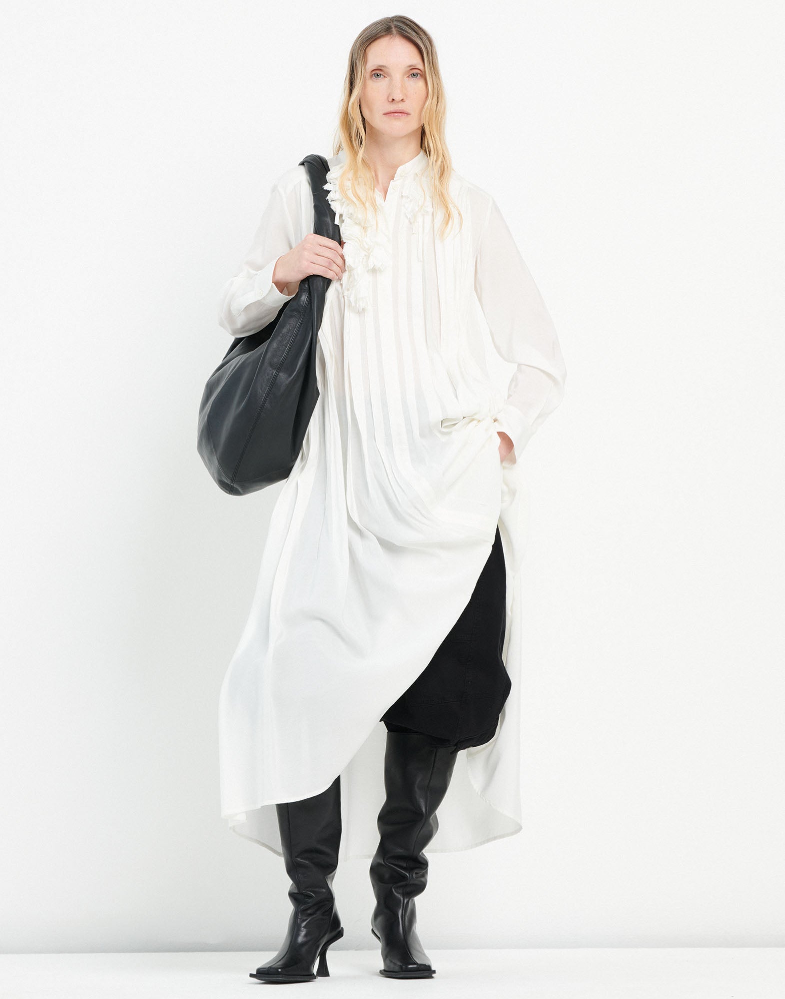 Front view: A woman wears the OUT RUN trousers with a wide silhouette and striking topstitching under a long white dress and knee-high black boots.