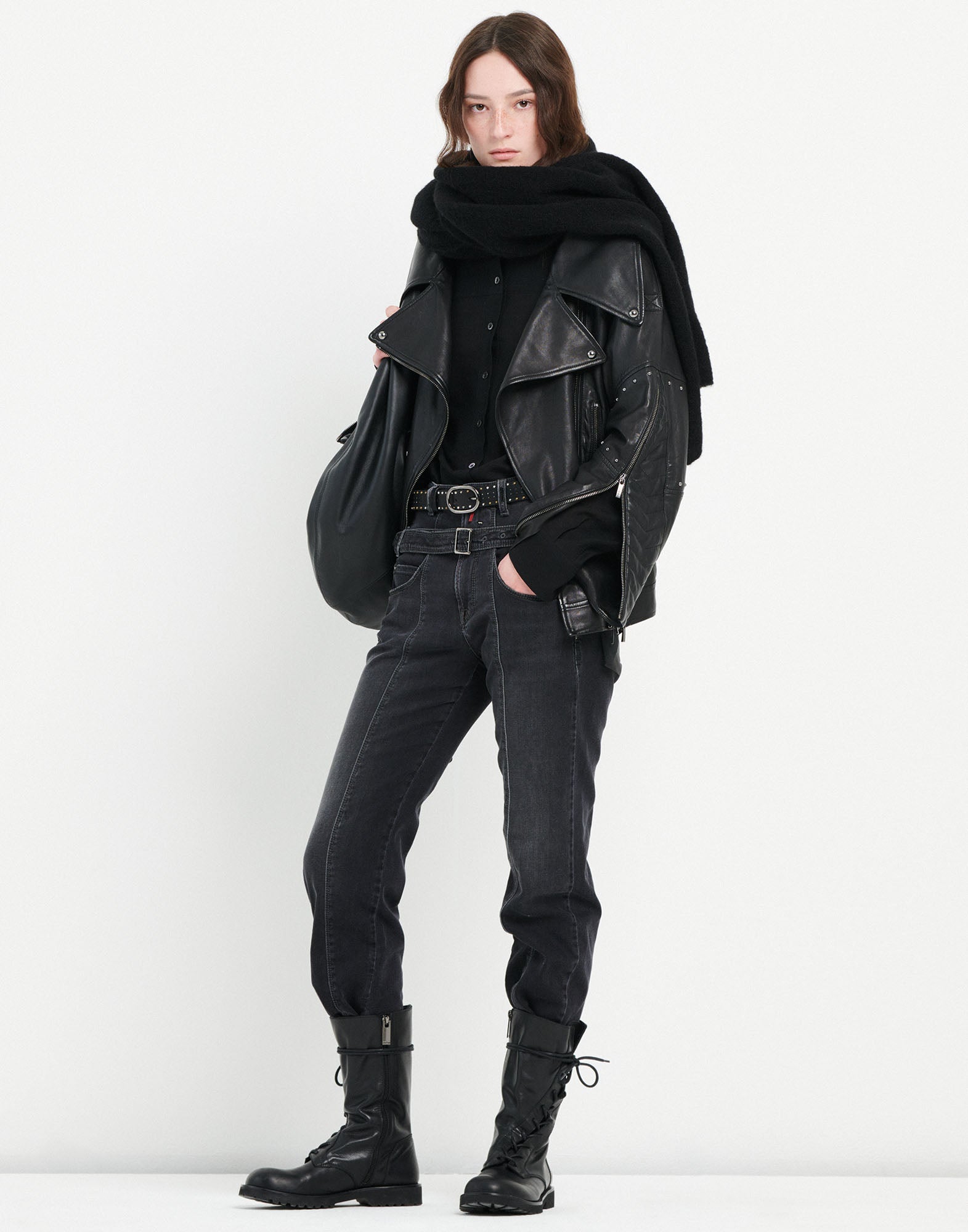 Frontalansicht einer Person, die die schwarze Slim-Fit-Jeans FAIR-PLAY mit abnehmbarem doppelten Gürtel trägt. Kombiniert mit Lederjacke, Schal und Boots.