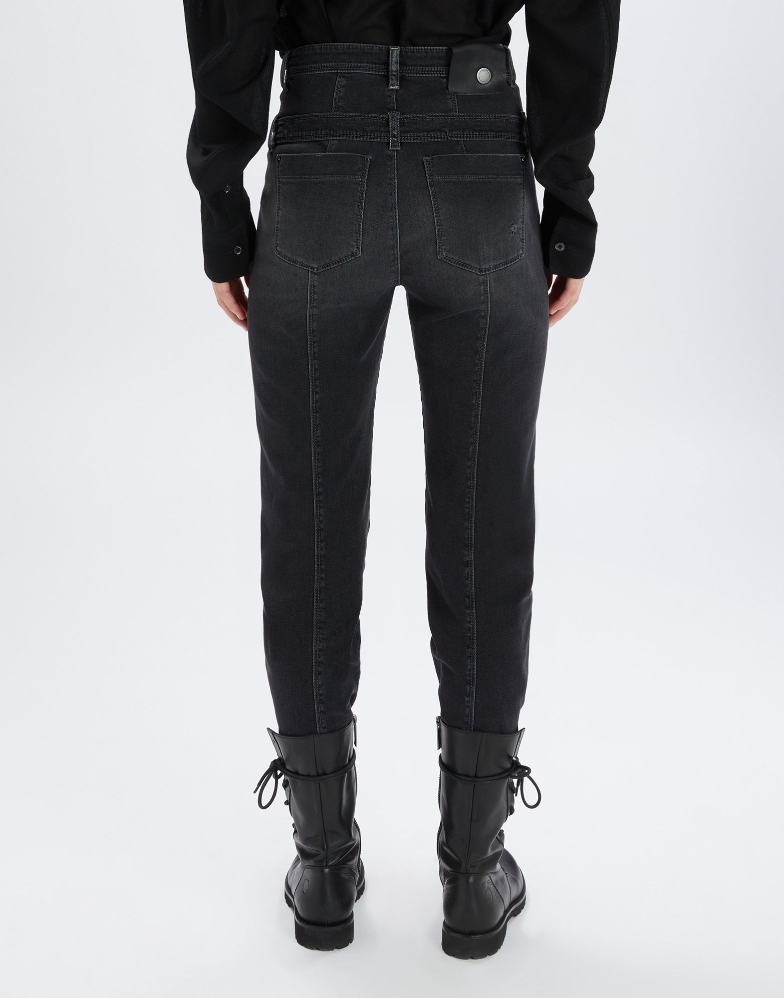 Rückansicht einer Person, die die schwarze Slim-Fit-Jeans FAIR-PLAY mit doppeltem, abnehmbarem Gürtel und sichtbaren Gesäßtaschen trägt. Die Jeans reicht bis zu den Knöcheln und wird mit schwarzen Stiefeln kombiniert.