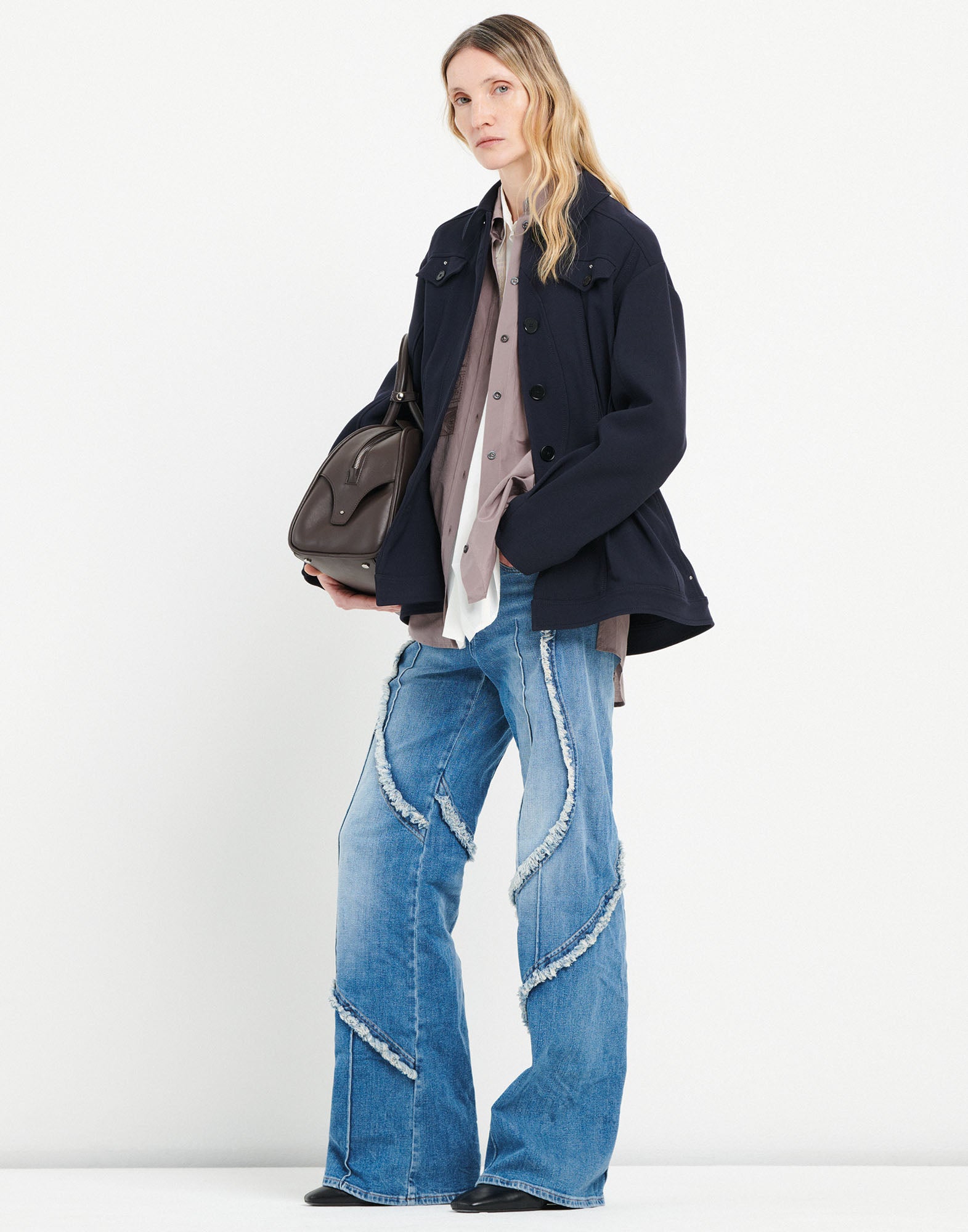 Frontalansicht: Modell trägt INDULGE Jeans mit weitem Bein, offenen, ausgefransten Kanten und sichtbaren Nähten. Heller Denim-Farbton, kombiniert mit dunkler Jacke.