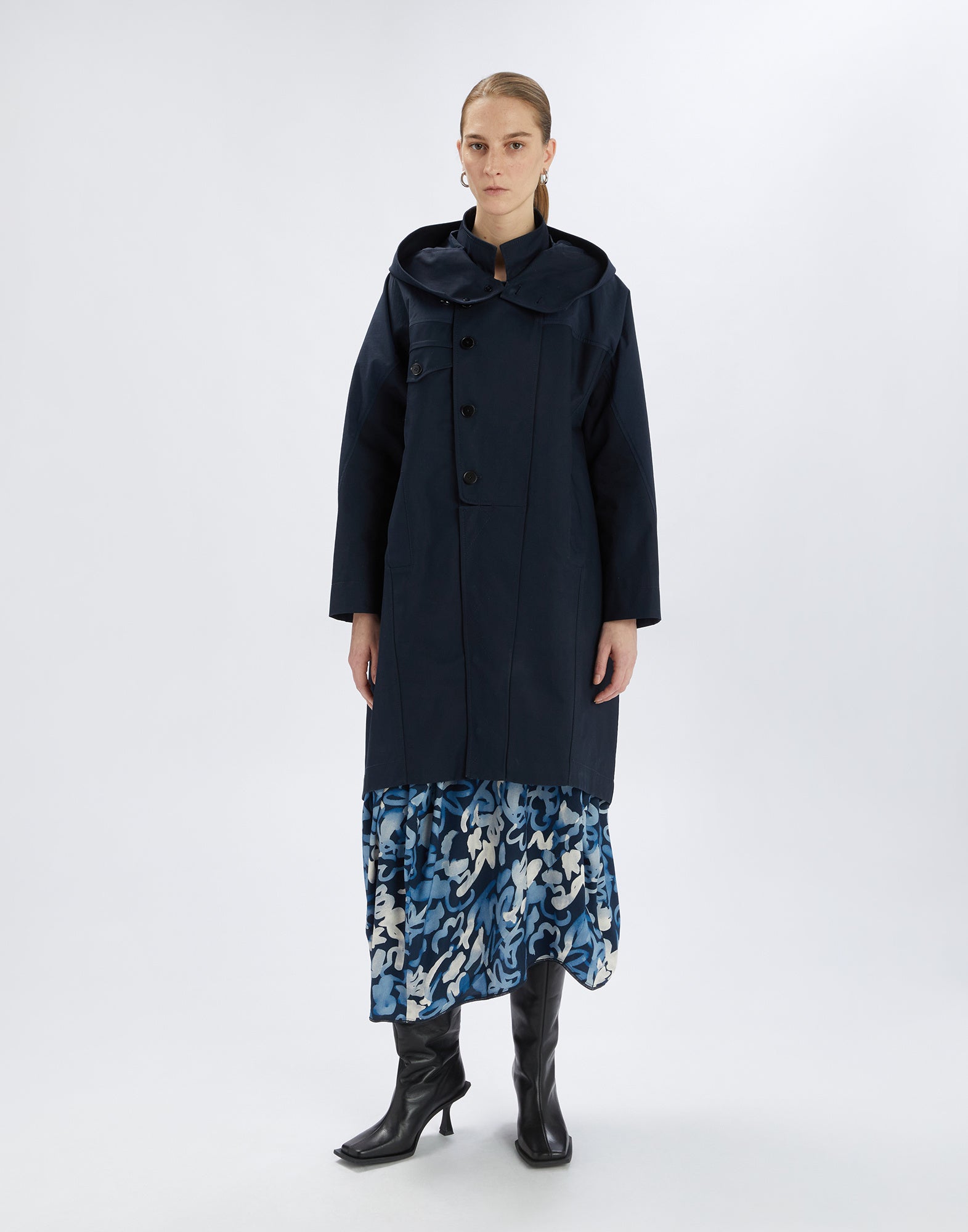 Frontalansicht: Eine Person trägt den dunkelblauen Parka UPGRADE mit hoher Knopfleiste, integrierter Kapuze und glatter Oberfläche über einem gemusterten Kleid.