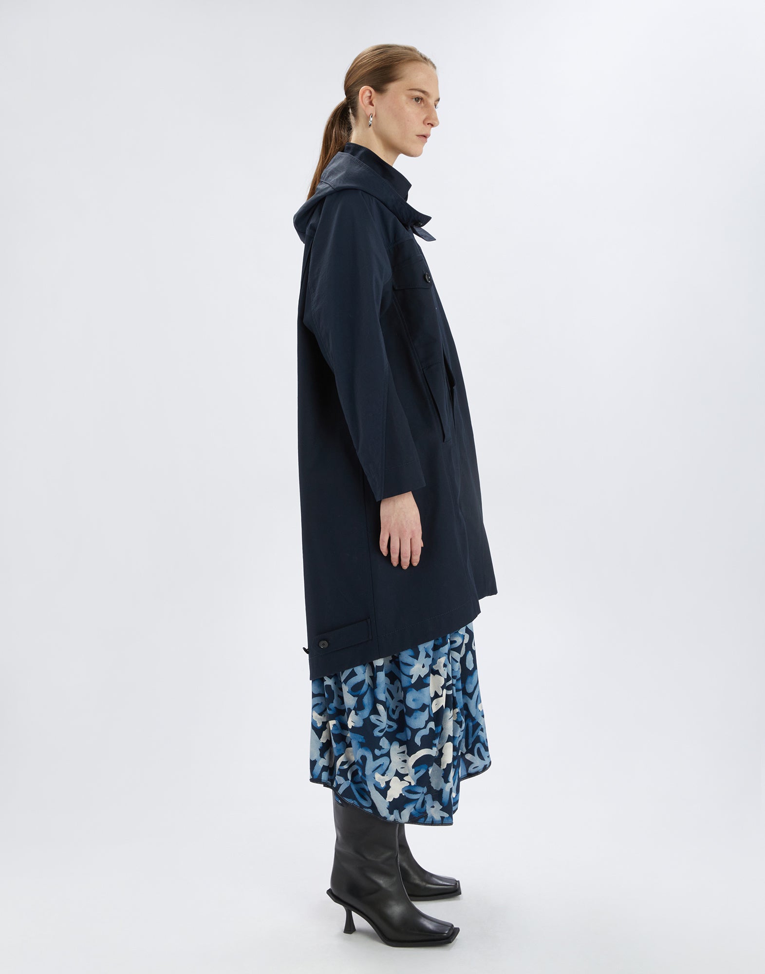 Seitenansicht vom UPGRADE Parka: sportlicher, dunkelblauer Mantel mit Kapuze, hohem Kragen, Knöpfen und Taschen. Das Modell trägt darunter einen blauen gemusterten Rock.