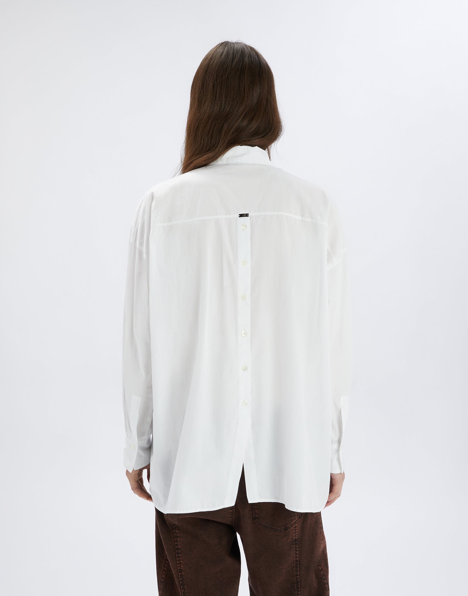 Rückansicht einer Person im weißen DEADLINE Hemd mit zentralem Knopfverschluss am Rücken, lockerer Passform und langem Ärmel, leicht über brauner Hose fallend.