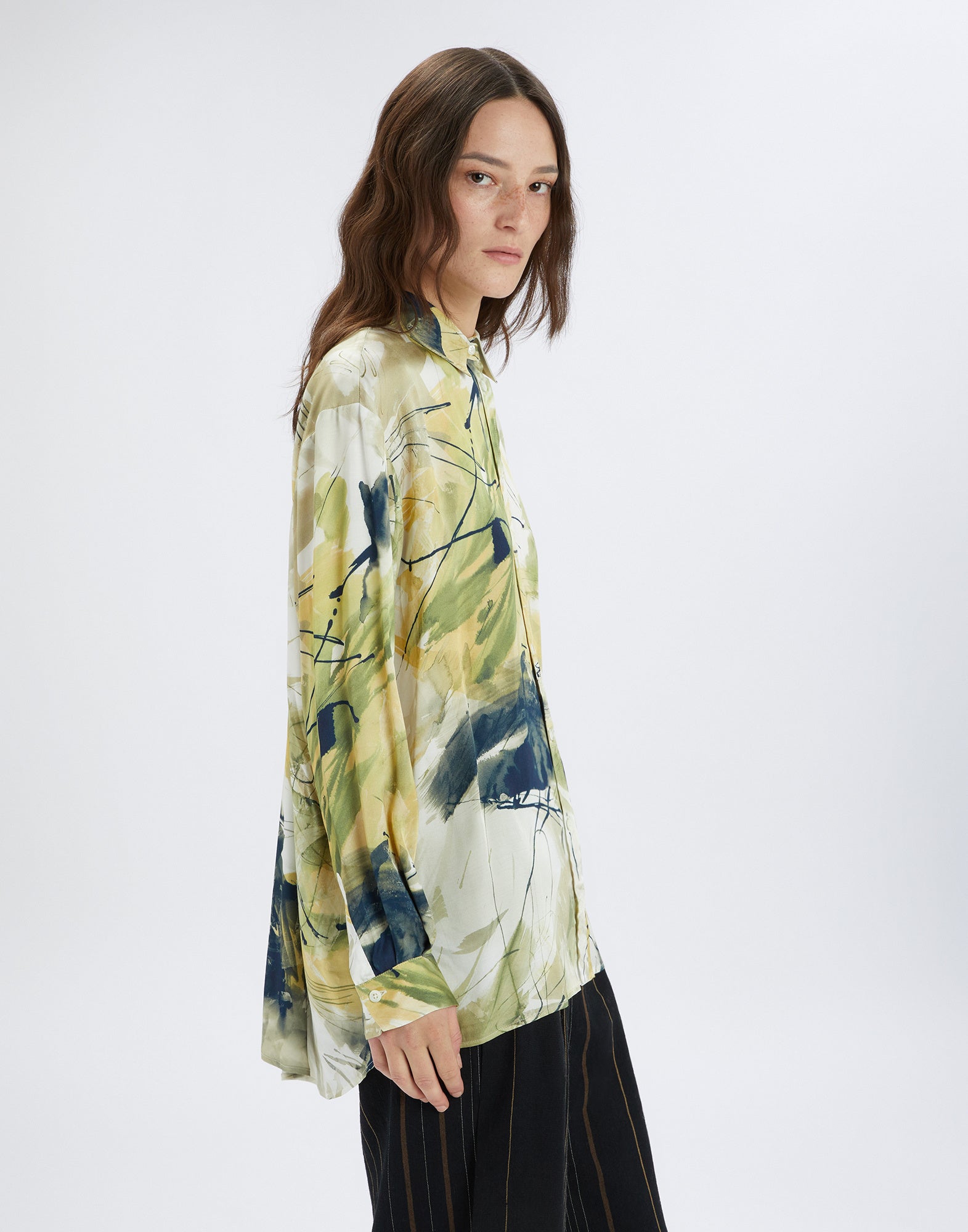 Side view: A model wears the KINFOLK shirt with an abstract, painterly pattern in yellow, green, and cream tones. Lightweight fabric, relaxed fit.