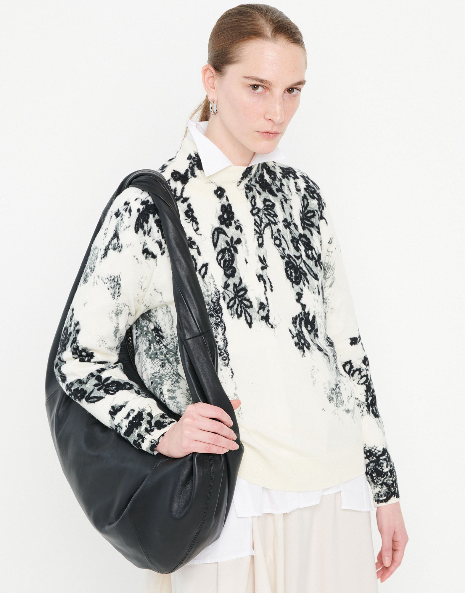 Frontalansicht: Person trägt den RAMSEY Merinowollpullover mit schwarzem floralen Muster auf cremefarbenem Grund, Stehkragen und Rippenbündchen, kombiniert mit großer schwarzer Tasche.