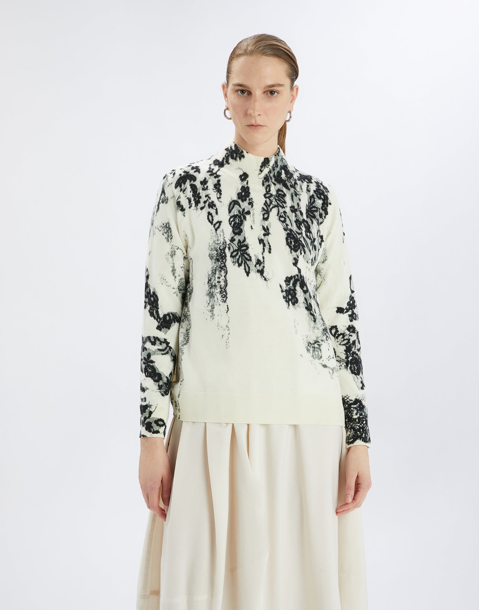 Frontalansicht: Modell trägt den RAMSEY Pullover mit schwarzem floralen Muster auf cremefarbenem Hintergrund, Stehkragen und Rippenbündchen, kombiniert mit hellem Rock.