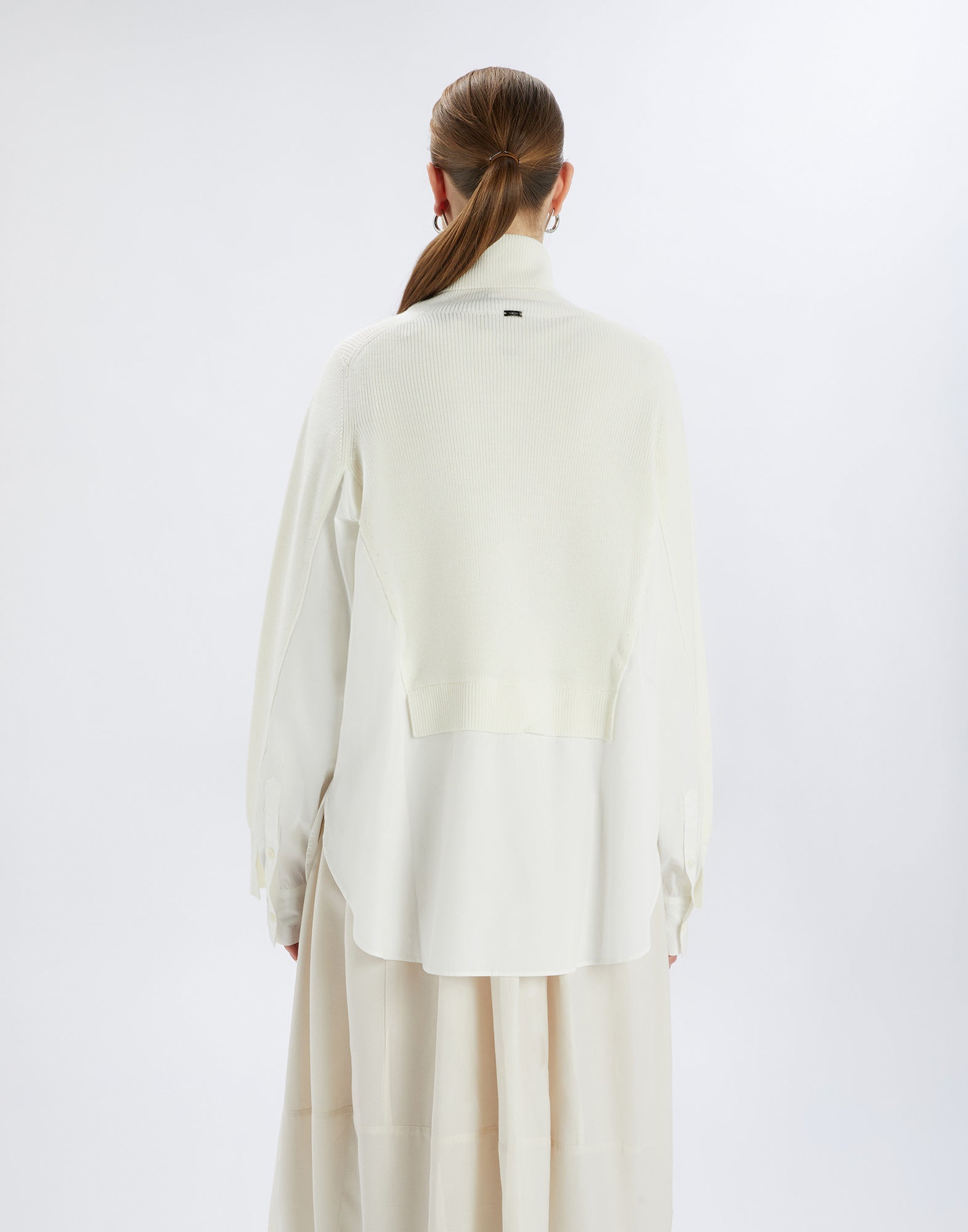 Rückansicht des Pullis HEART-BIT, getragen von einer Frau mit zusammengebundenem Haar. Der weiße Woll- und Baumwollpullover ahmt einen Lagenlook nach.