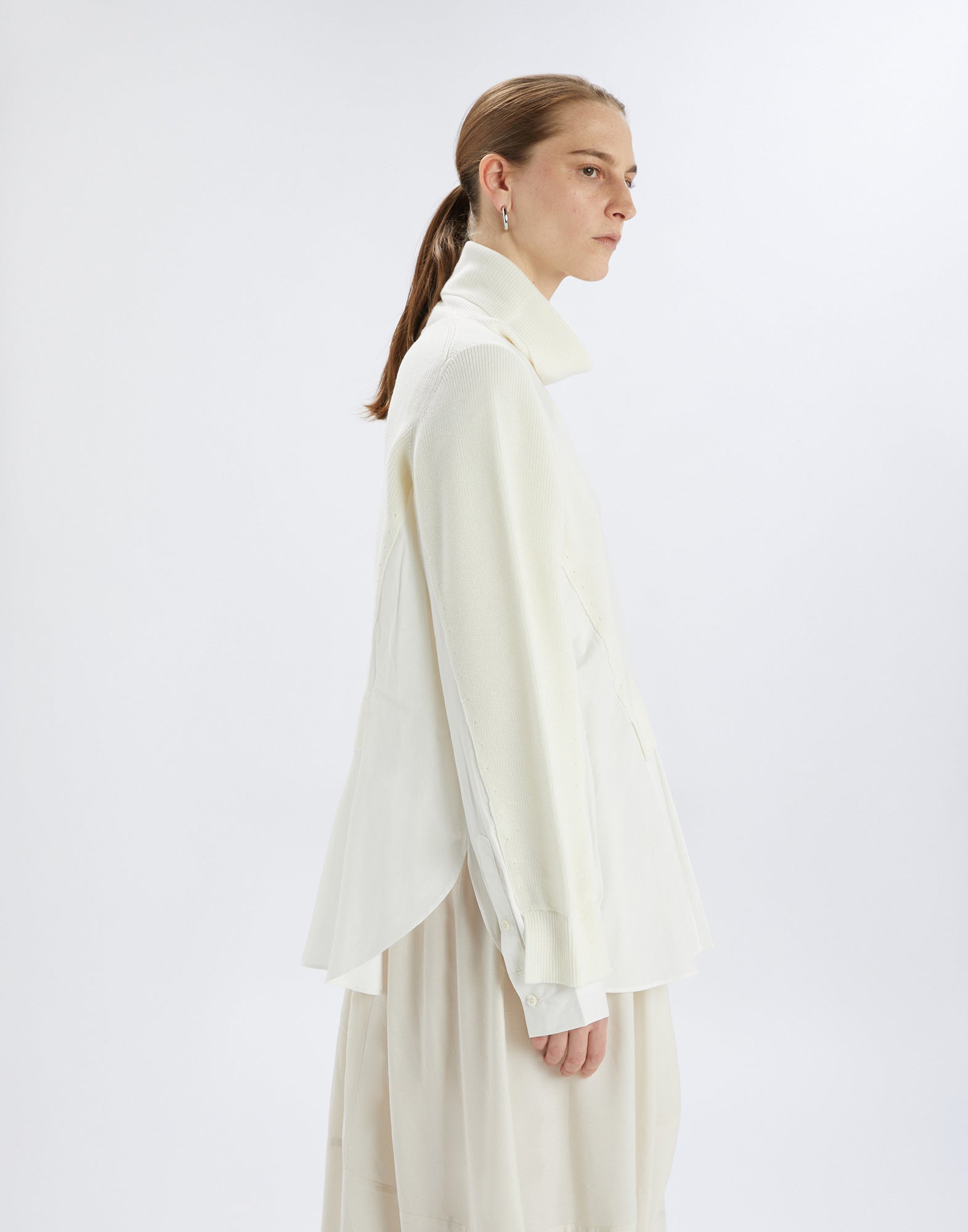 Seitenansicht eines Modells, das den HEART-BIT Pullover in Cremeweiß trägt. Hoher Kragen, lange Ärmel, kombinierte Wolle und Baumwollpopeline mit schichtartigem Effekt.