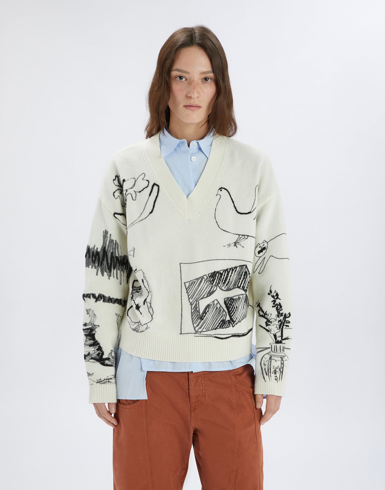 Frontalansicht eines Modells, das den Pulli APLOMB trägt. Der Pullover hat eine Boxy-Passform, V-Ausschnitt und zeigt stilisierte, schwarz gedruckte und gestickte Designs auf hellem Wollstoff.