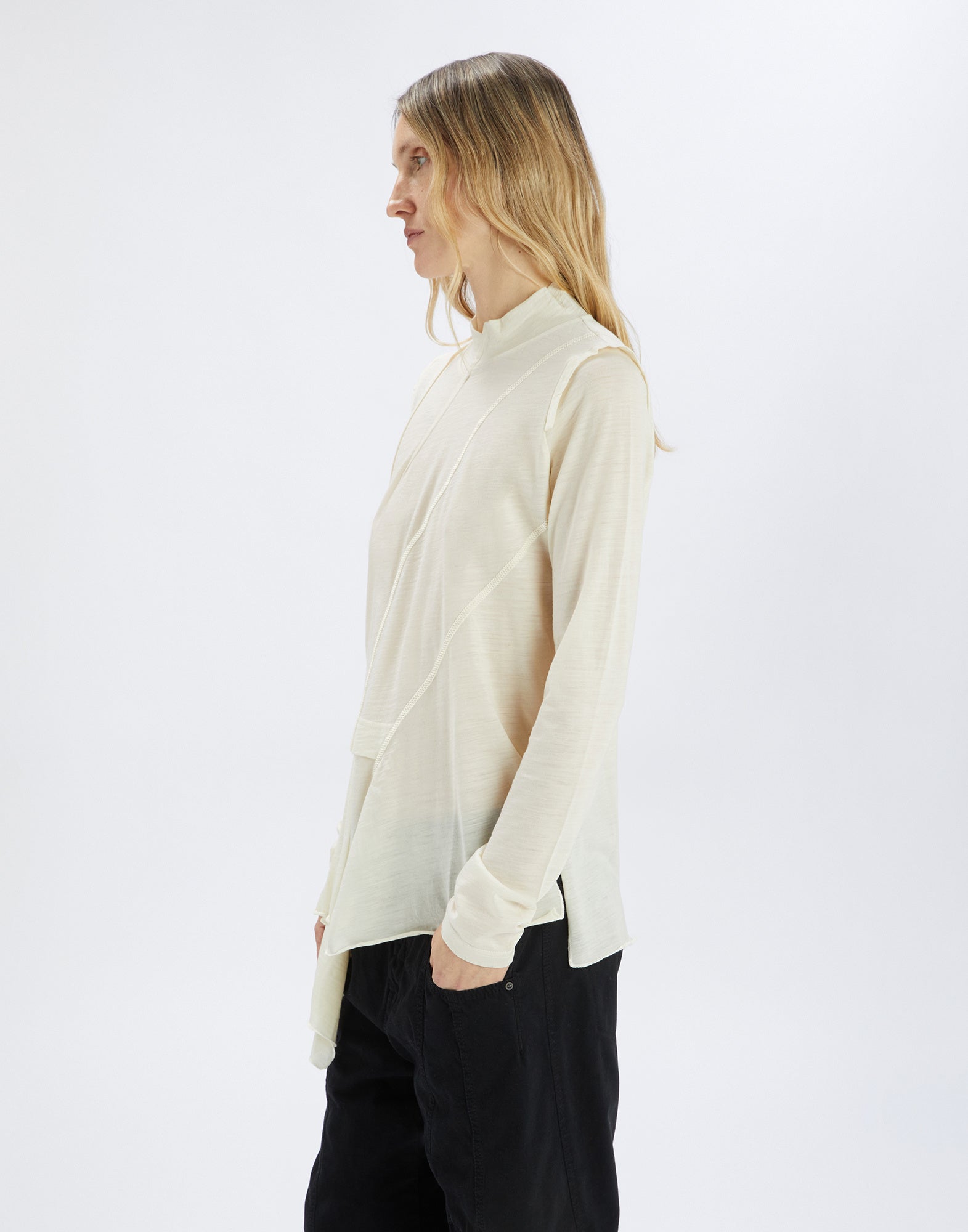 Seitenansicht einer Person, die den cremefarbenen MATELOT Pullover trägt. Locker geschnittenes, langärmliges Modell mit asymmetrischem Saum und Stehkragen.