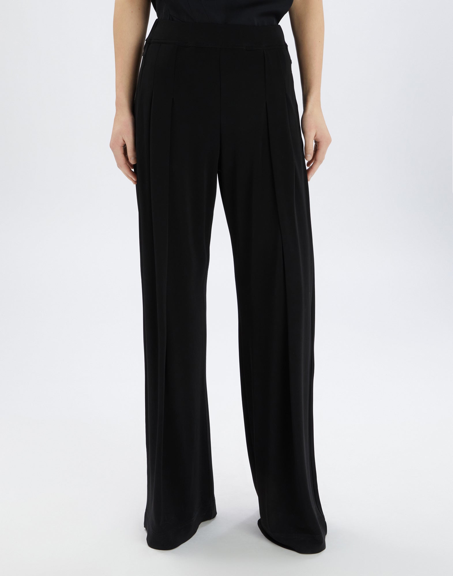 Front view: A person wears SLIP-OVER pants in black viscose jersey with wide legs, front pleats, and an elastic waistband for an elegant pajama style.