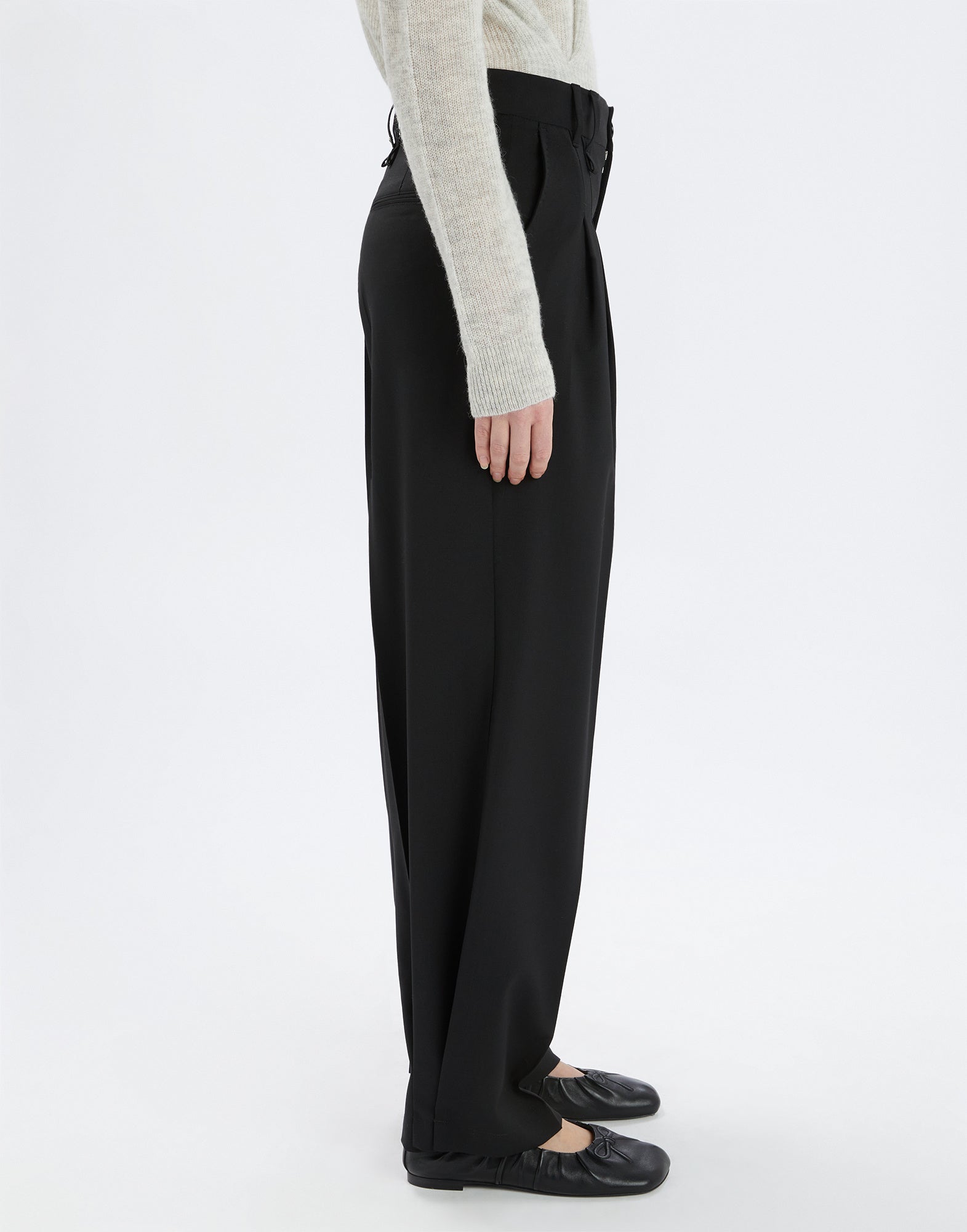 Side view of the RELY pants in extra-fine black wool, relaxed fit with front pleats and darted hem, worn by a person in a light-colored top.