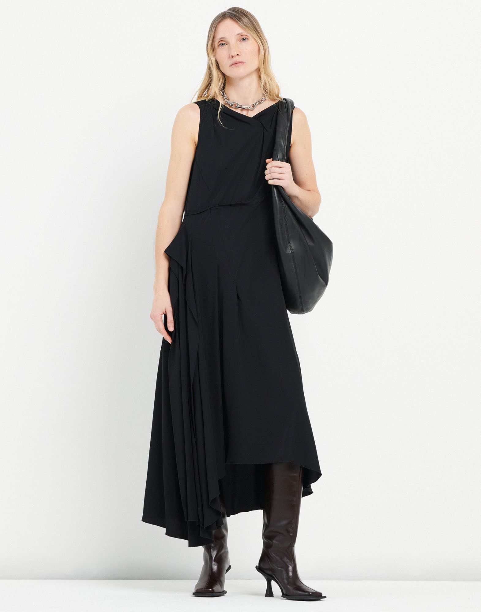 Frontalansicht: SOUND OFF Kleid mit asymmetrischem Schnitt, ärmellos, in Schwarz. Weich fallender Stoff an der Hüfte, getragen mit hohen dunkelbraunen Stiefeln und großer schwarzer Tasche.