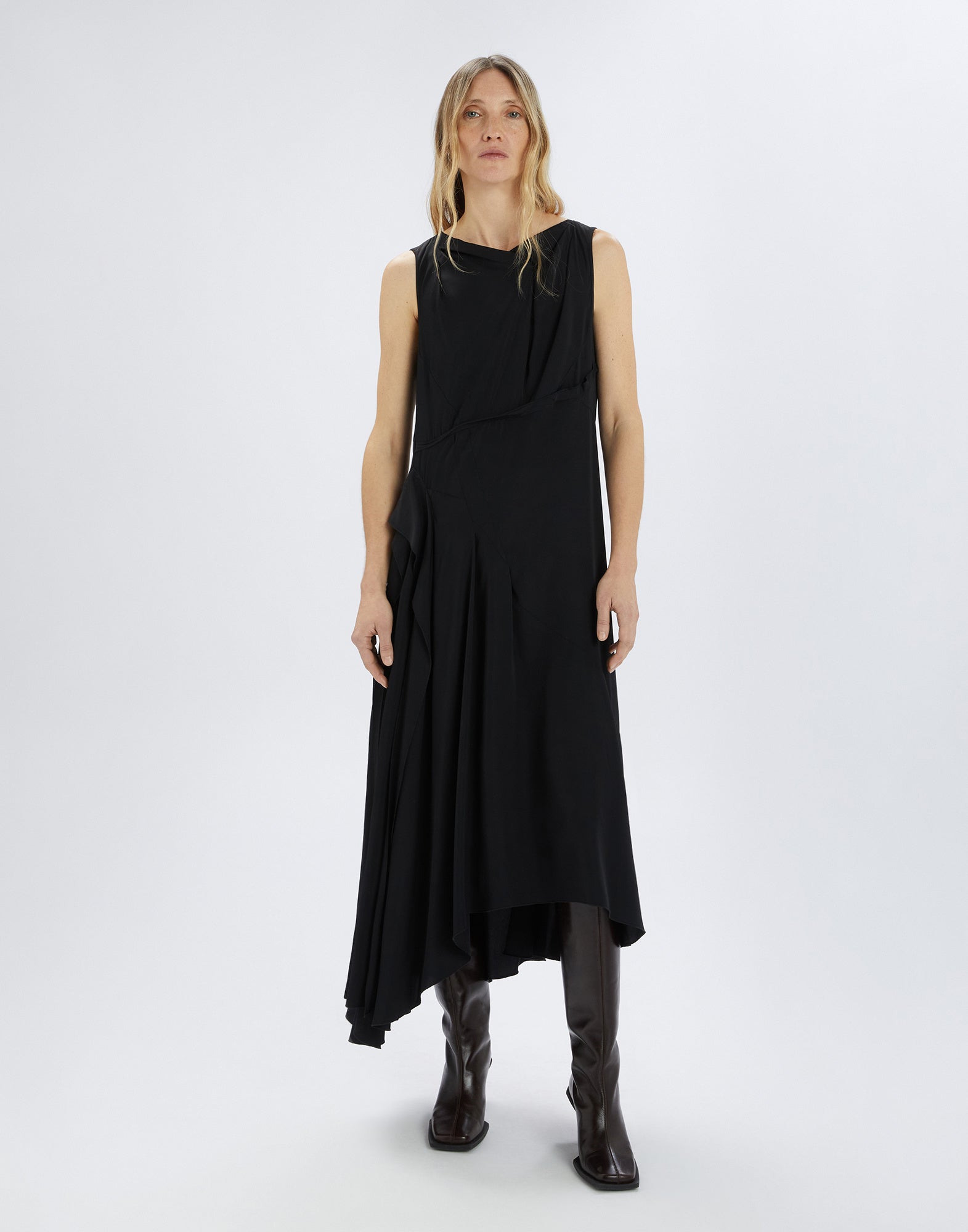 Front view of a woman wearing the SOUND OFF sleeveless dress with an asymmetric cut and side draping. The fabric flows softly over knee-high dark boots.