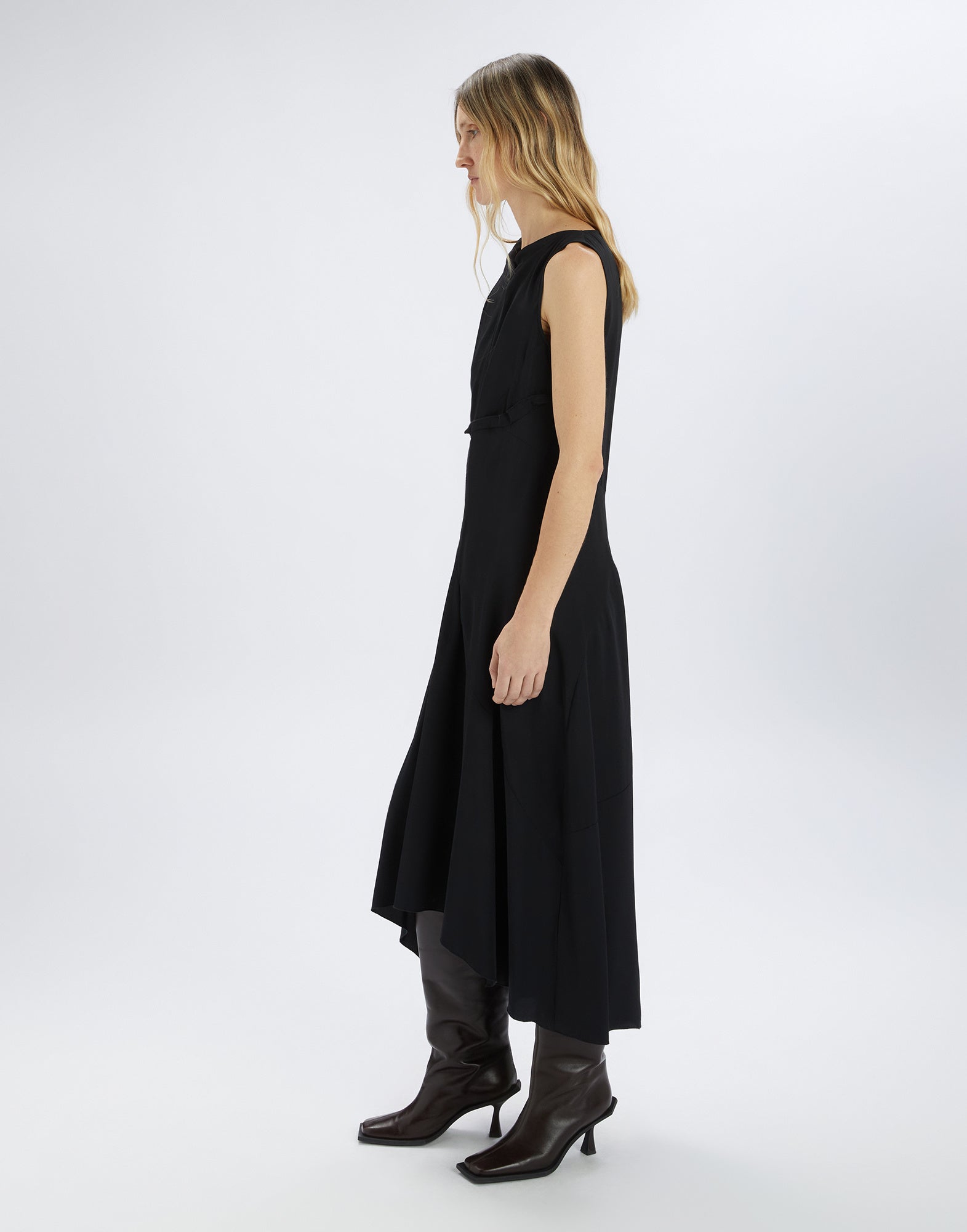 Side view of a model wearing the SOUND OFF sleeveless black dress with asymmetric hem and side draping, paired with dark boots.