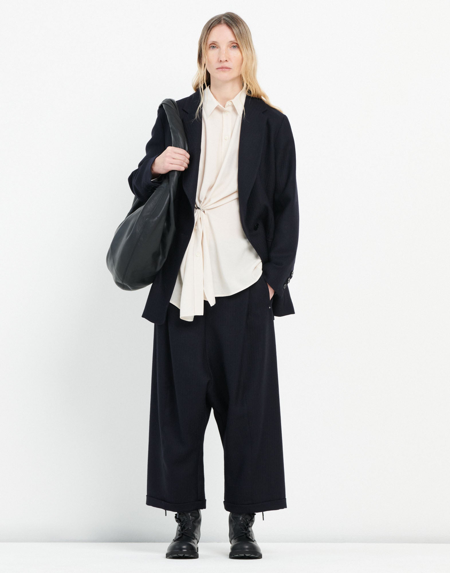 Front view: A woman wears the SKYLARK blouse with elegant pleating through a central ring, loose silhouette and classic button placket, paired with a dark blazer and wide-leg trousers.