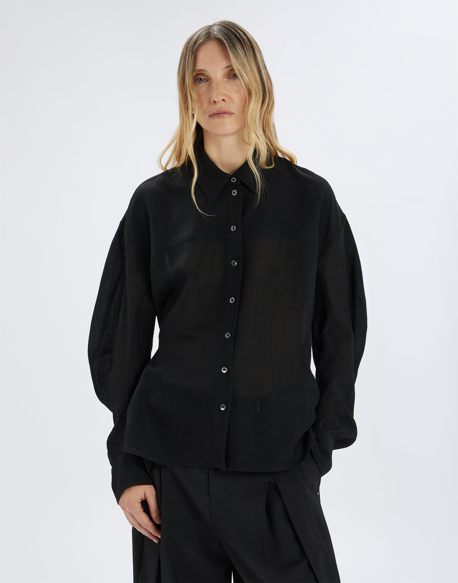 Front view: A woman wears the black SCOOT blouse with long puff sleeves, front button placket, and classic notched collar made from nearly transparent extra-fine wool gauze.