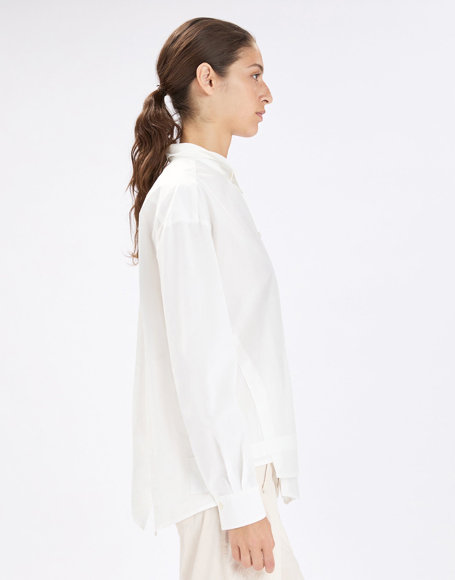 Side view: person wearing the relaxed-fit white SAWYER shirt with classic collar, long sleeves, and asymmetrical, longer-back hem over light pants against a neutral background.