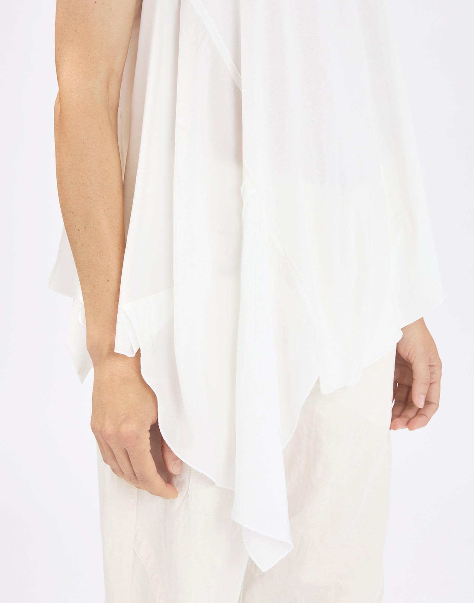 Detail view: person wearing the sleeveless COMPASS top in white, front view focused on the right side with softly draping ruffle and irregular, pointed hem over light-colored trousers.