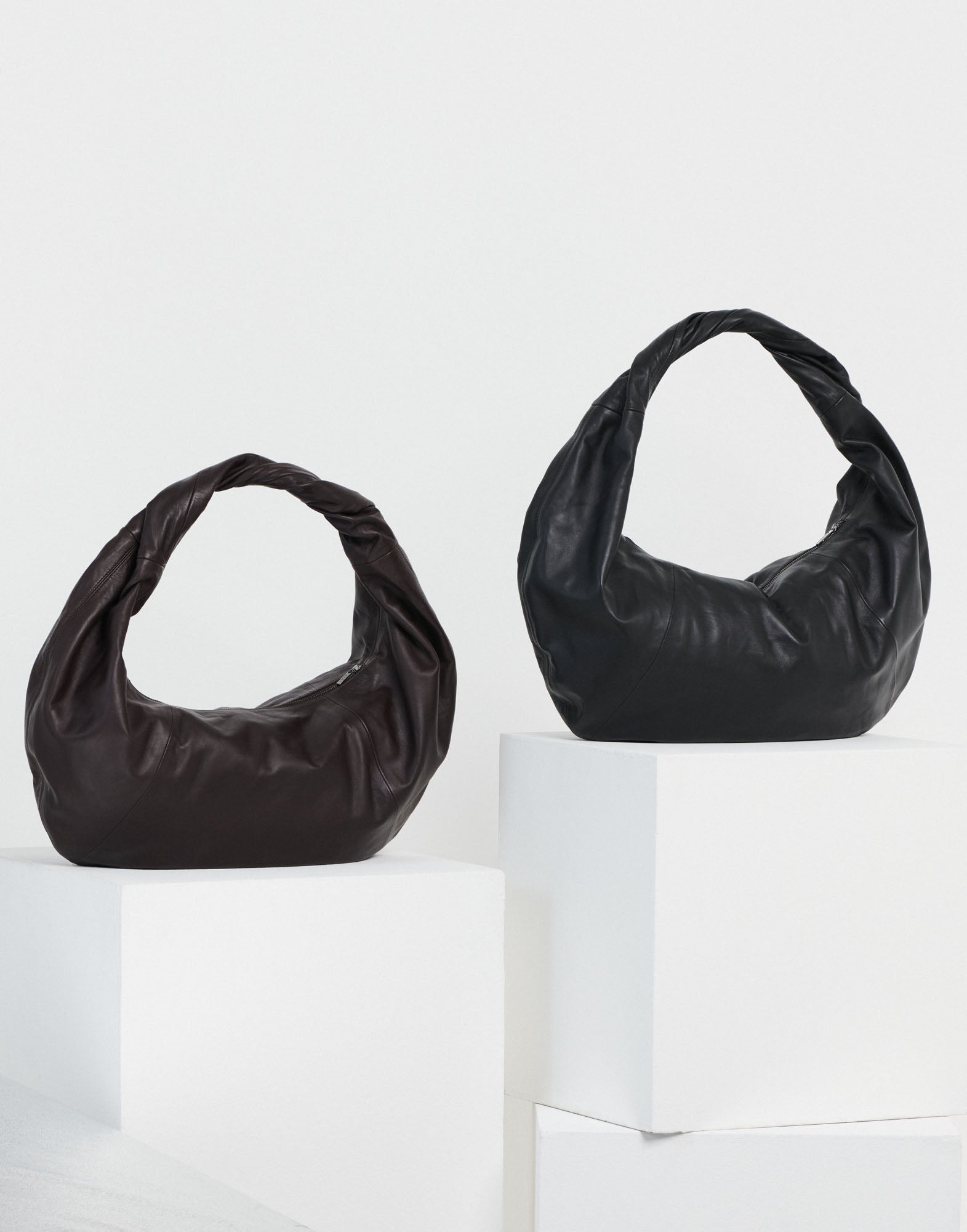 Frontalansicht von zwei REFLEX Hobo-Taschen in Braun und Schwarz mit geflochtenem Henkel aus weichem Nappaleder, ausgestellt auf weißen Podesten.