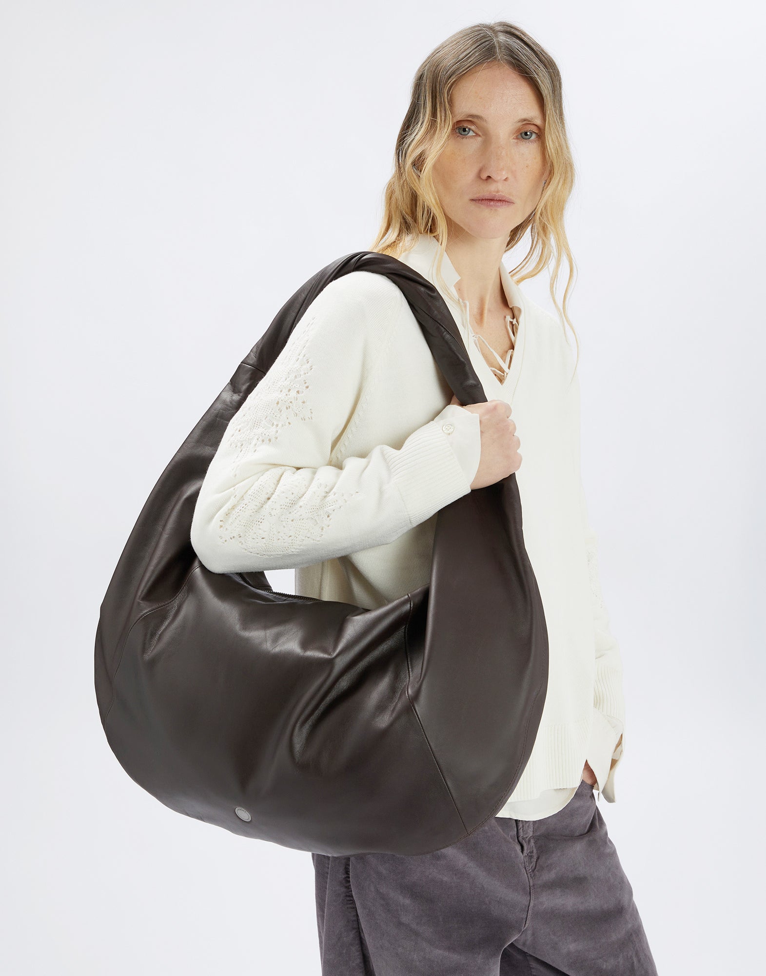 Front view: A woman carries the large REFLEX hobo bag in soft dark nappa leather on her shoulder. The bag features a braided handle and a rounded shape.