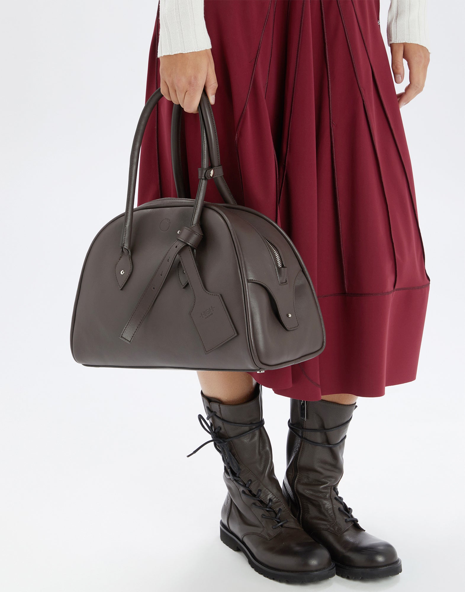Front view: A person holds the BELUGA Bowling Bag in dark brown calf leather with reinforced handles, metal feet, and decorative leather tag in hand.