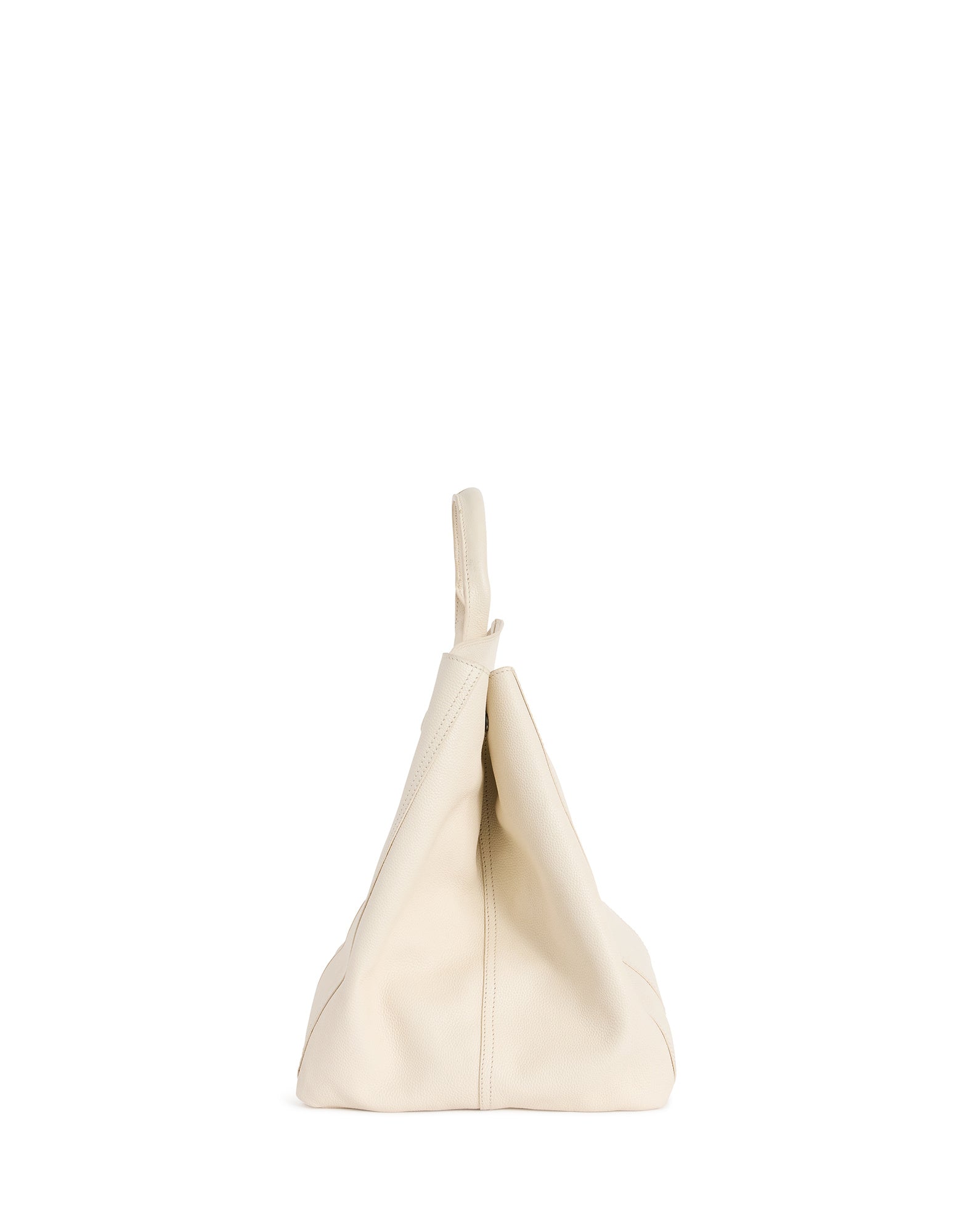 Seitenansicht: Die Lederhandtasche CUTE steht leicht gefaltet, weiches ungeformtes beigefarbenes Leder mit sichtbaren Nähten und nach oben ragendem doppeltem Griff vor weißem Hintergrund.