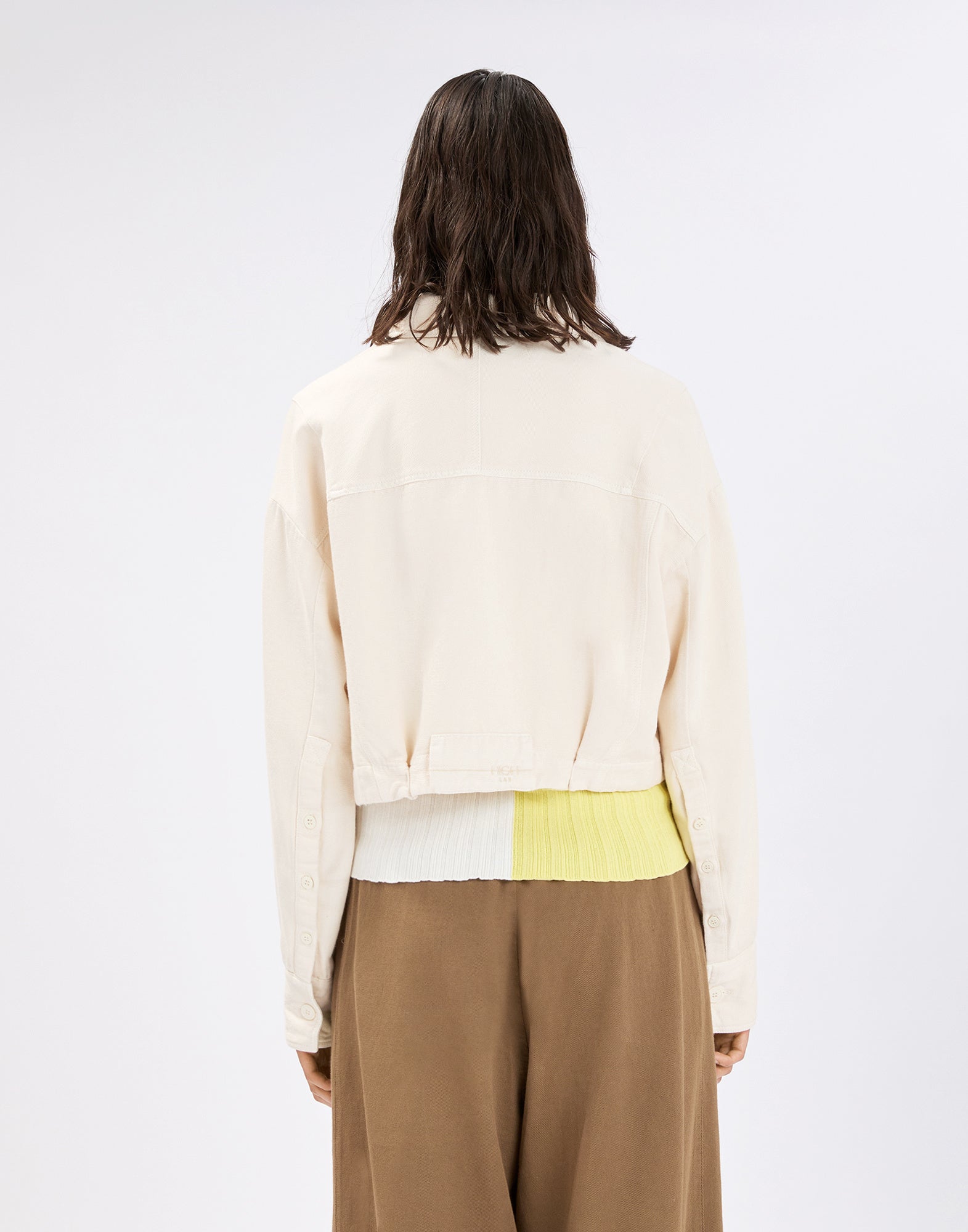 Back view: person wearing the cropped cream DEBUT cotton-linen jacket with button plackets on the sleeves, over a two-tone knitted waistband and wide brown pants.