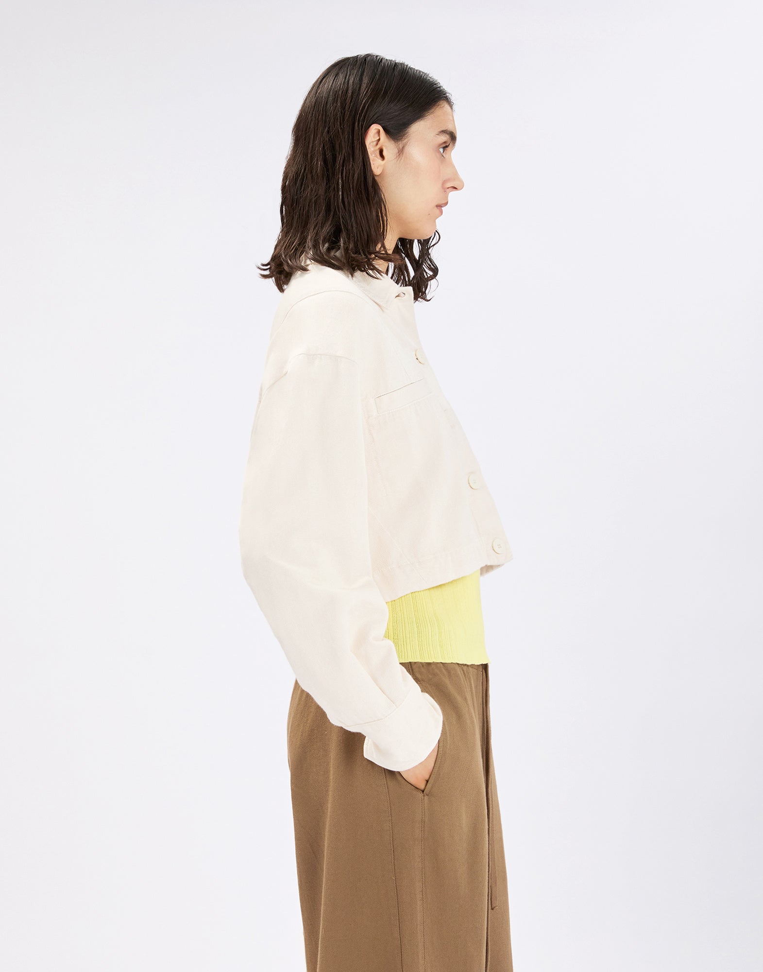 Side view: person wearing the slightly cropped, cream-colored DEBUT cotton-linen jacket with shirt collar and button placket, paired with a yellow top and brown pants against a neutral background.