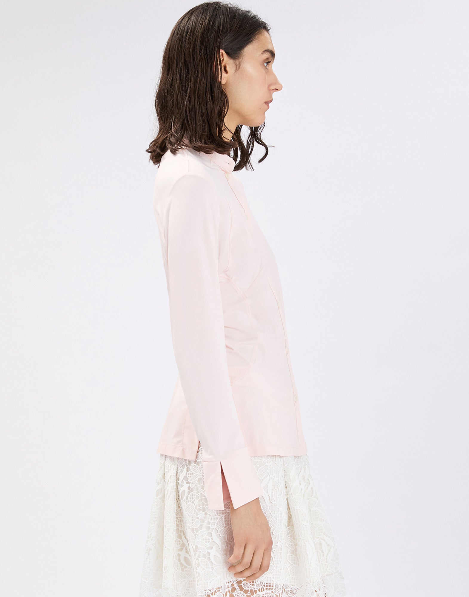 Side view: model wears the fitted INDEED shirt in light pink jersey with mandarin collar, long sleeves and tall cuff with slit, paired with a white lace skirt.