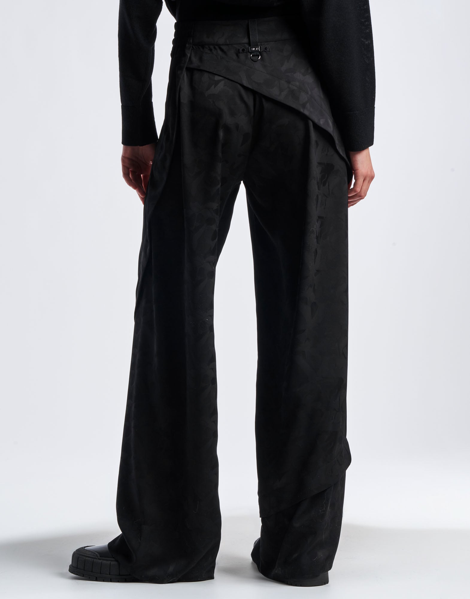 Rückansicht der BORDERLINE Hose mit weitem Bein aus schwarzem Funktionssatin mit Damast-Blumenmuster, breitem Saum und asymmetrischen, spiralförmigen Nähten.