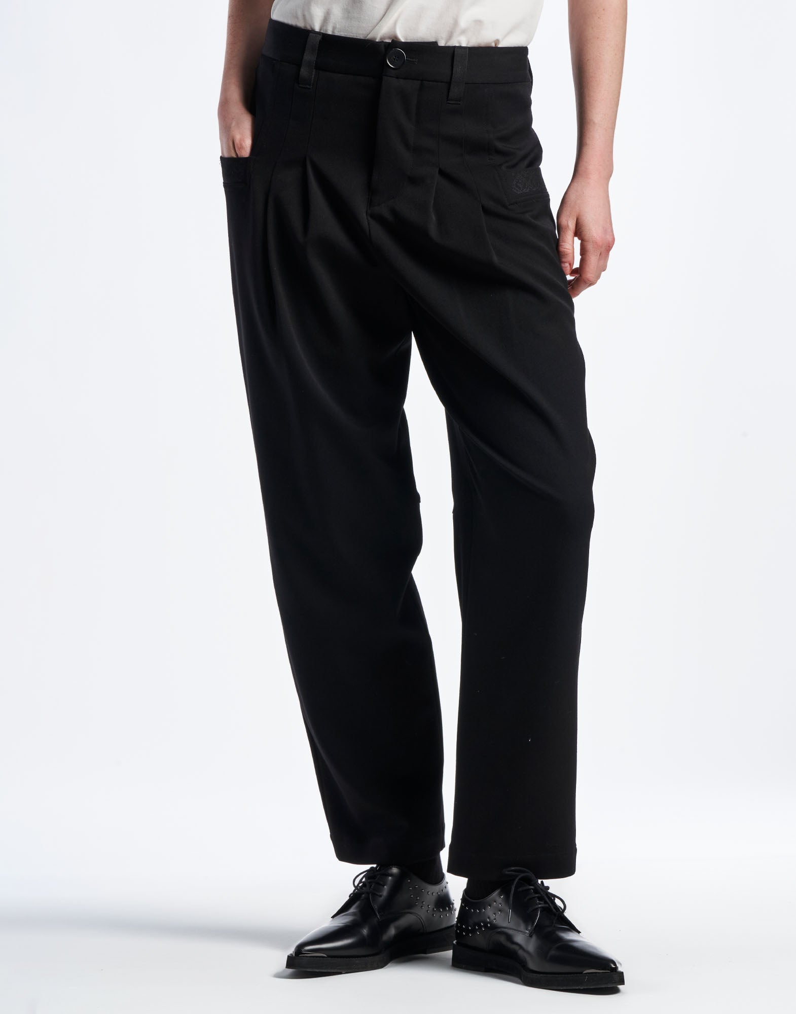 Frontalansicht der schwarzen Hose COMPLY aus weichem Funktionstwill, Taillenbund mit Gürtelschlaufen, Falten und geometrisch bestickten Paspeltaschen, konisch geschnitten.