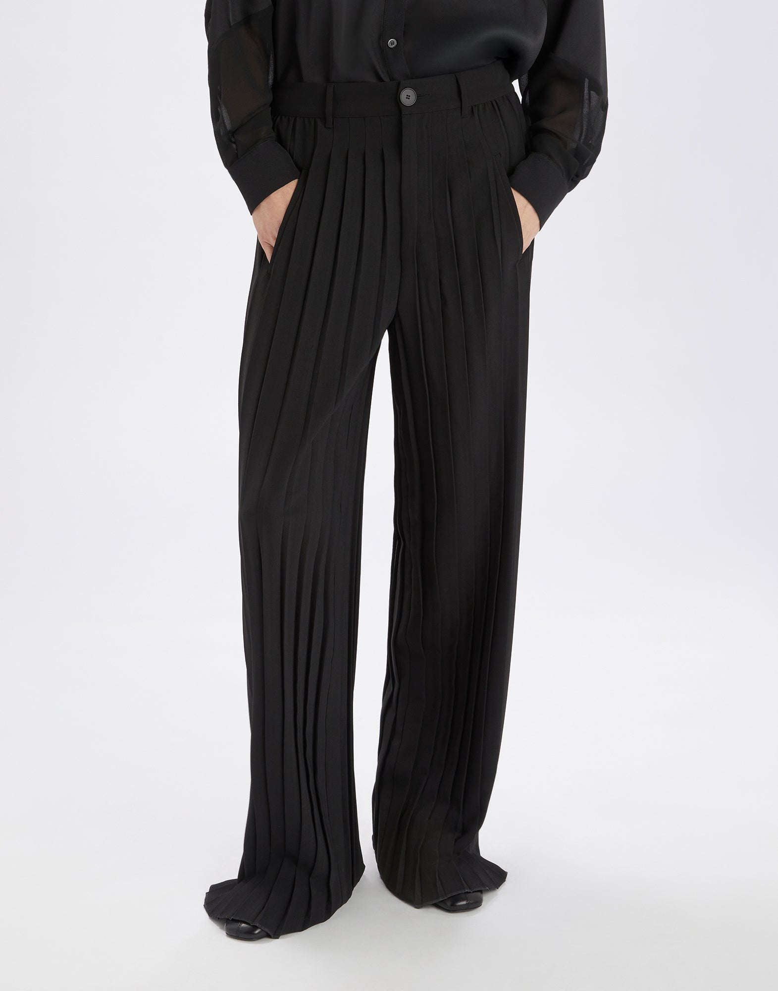 Front view: person wearing the DREW palazzo trousers in black, featuring prominent vertical pleats, straight leg, waistband with belt loops and button closure.