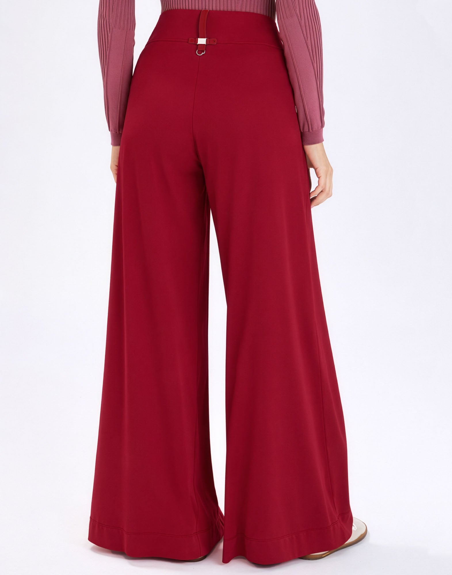 Rear view: model wearing the burgundy, very wide-leg DIMINISH pants with high waist, elastic waistband, and decorative back tab against a neutral background.