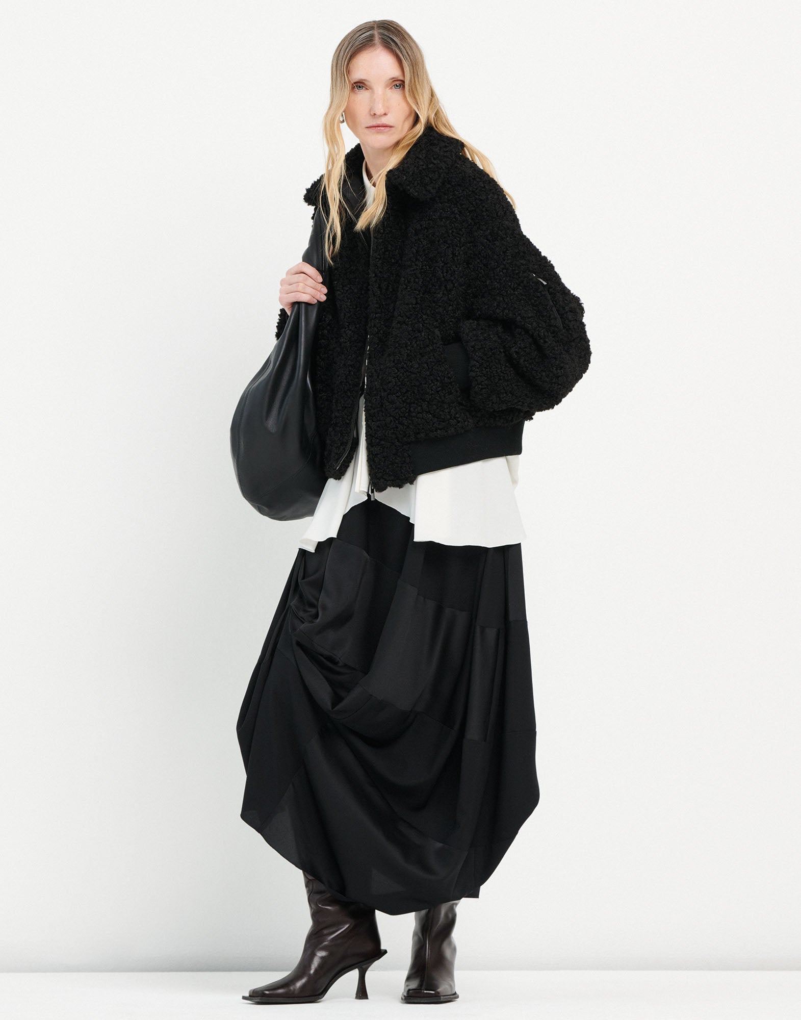 Frontalansicht: Eine Frau trägt den Rock GEEK aus schwarzem Tech-Satin mit asymmetrischer Drapierung, Midi-Länge und fließender Silhouette, kombiniert mit dunklen Stiefeln.