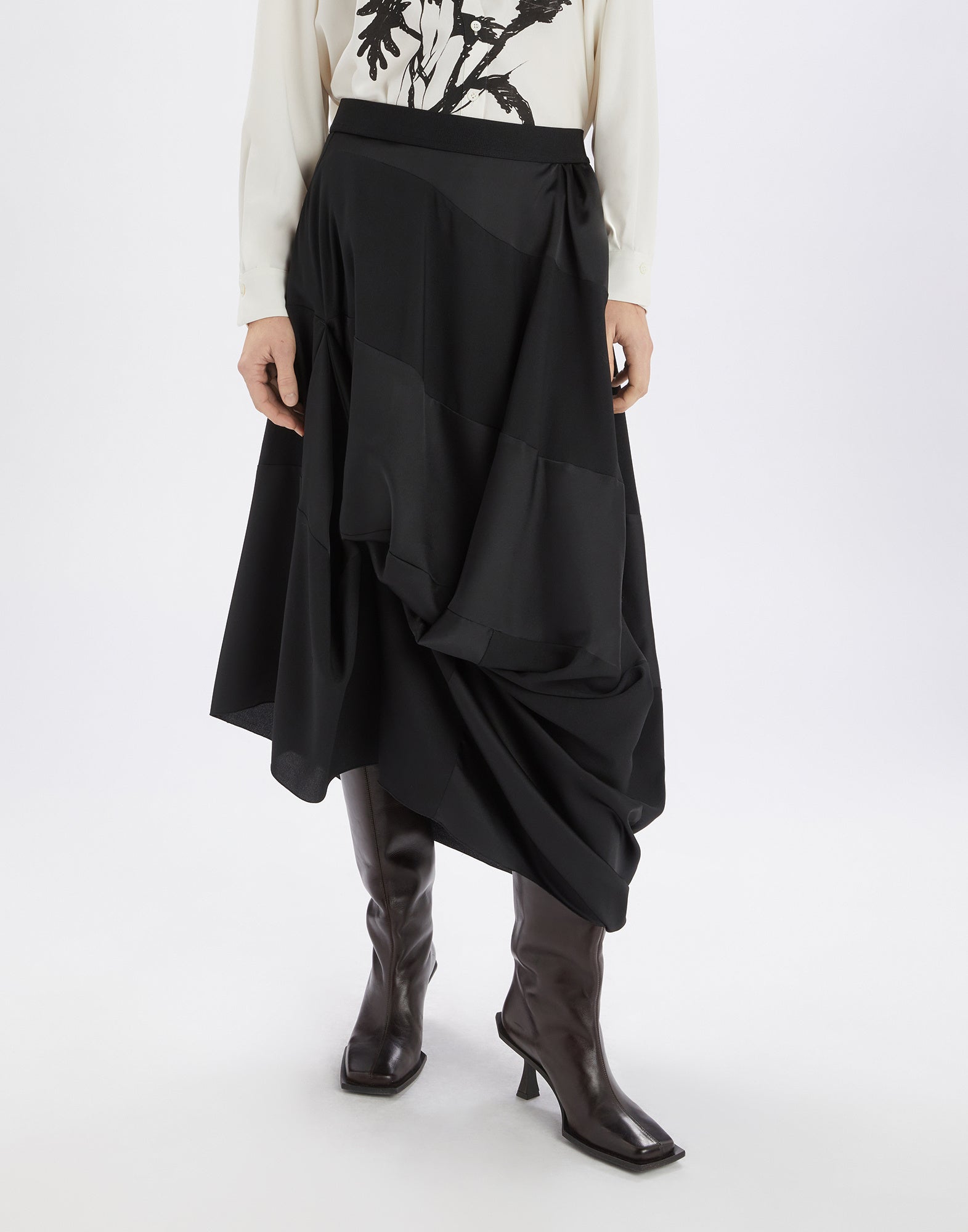 Front view of the GEEK skirt in black fluid tech satin with an asymmetric draped cut, elasticated waistband, and midi length, worn with knee-high boots.