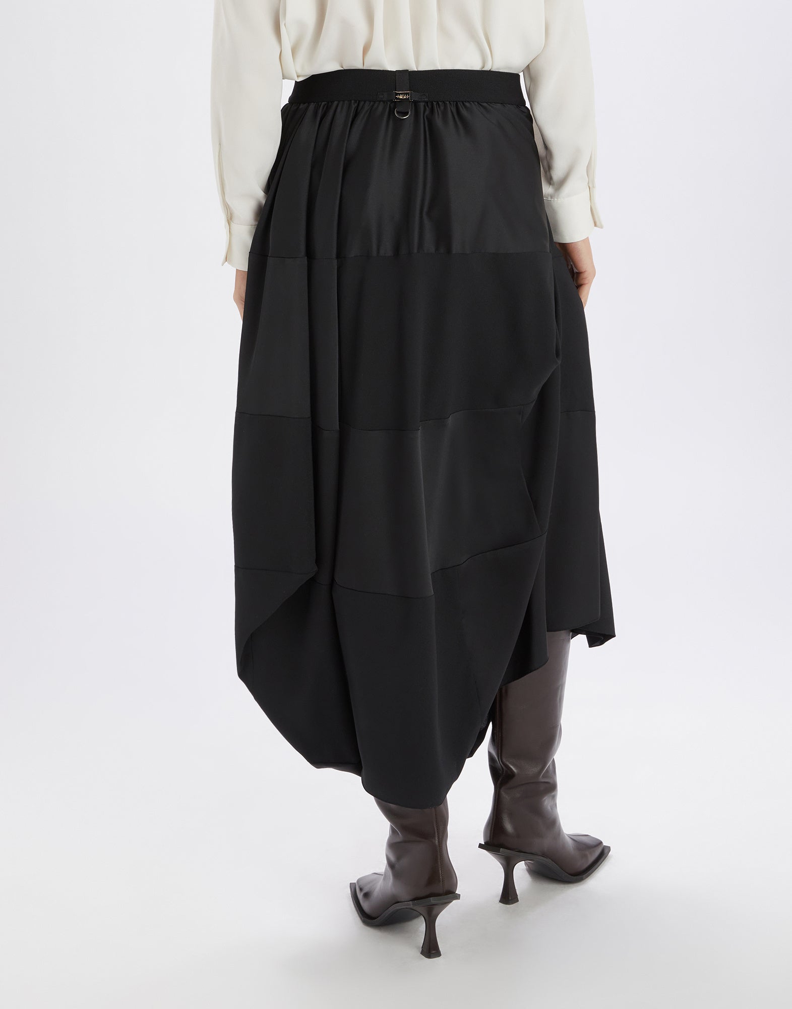 Back view of the GEEK skirt in black tech satin with elastic waistband and asymmetric, loose-falling hem at midi length. The wearer stands with brown boots.