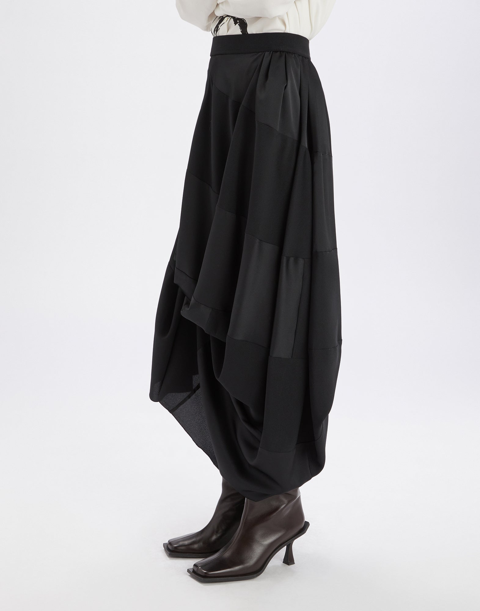 Side view of GEEK skirt in black fluid tech satin with asymmetric draping, elastic waistband, and midi length, worn with dark boots.