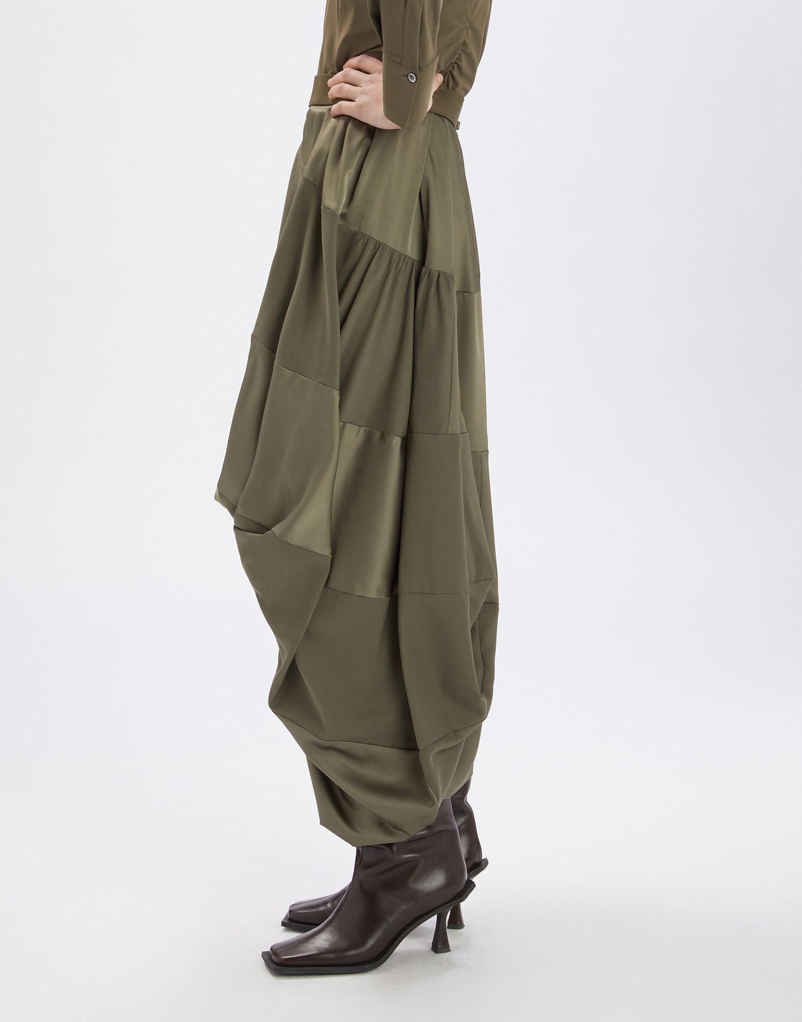 Seitenansicht des Rocks GEEK, aus olivgrünem Tech-Satin gefertigt, mit asymmetrischem, drapiertem Schnitt, Midi-Länge und elastischem Bund, getragen mit Stiefeletten.