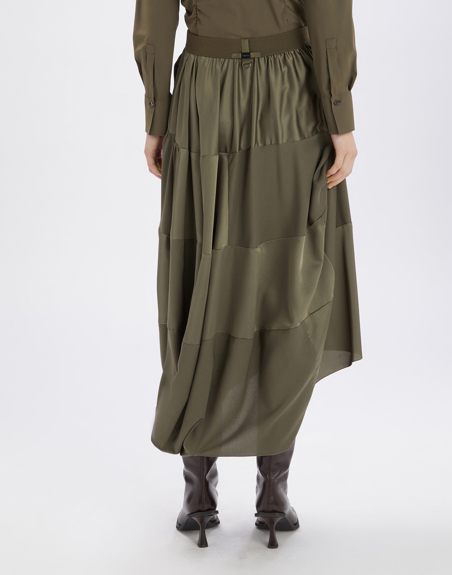 Rückansicht des GEEK-Rocks in olivgrün aus fließendem Tech-Satin, mit elastischem Bund, asymmetrischem Saum und Midi-Länge, getragen mit braunen Stiefeln.