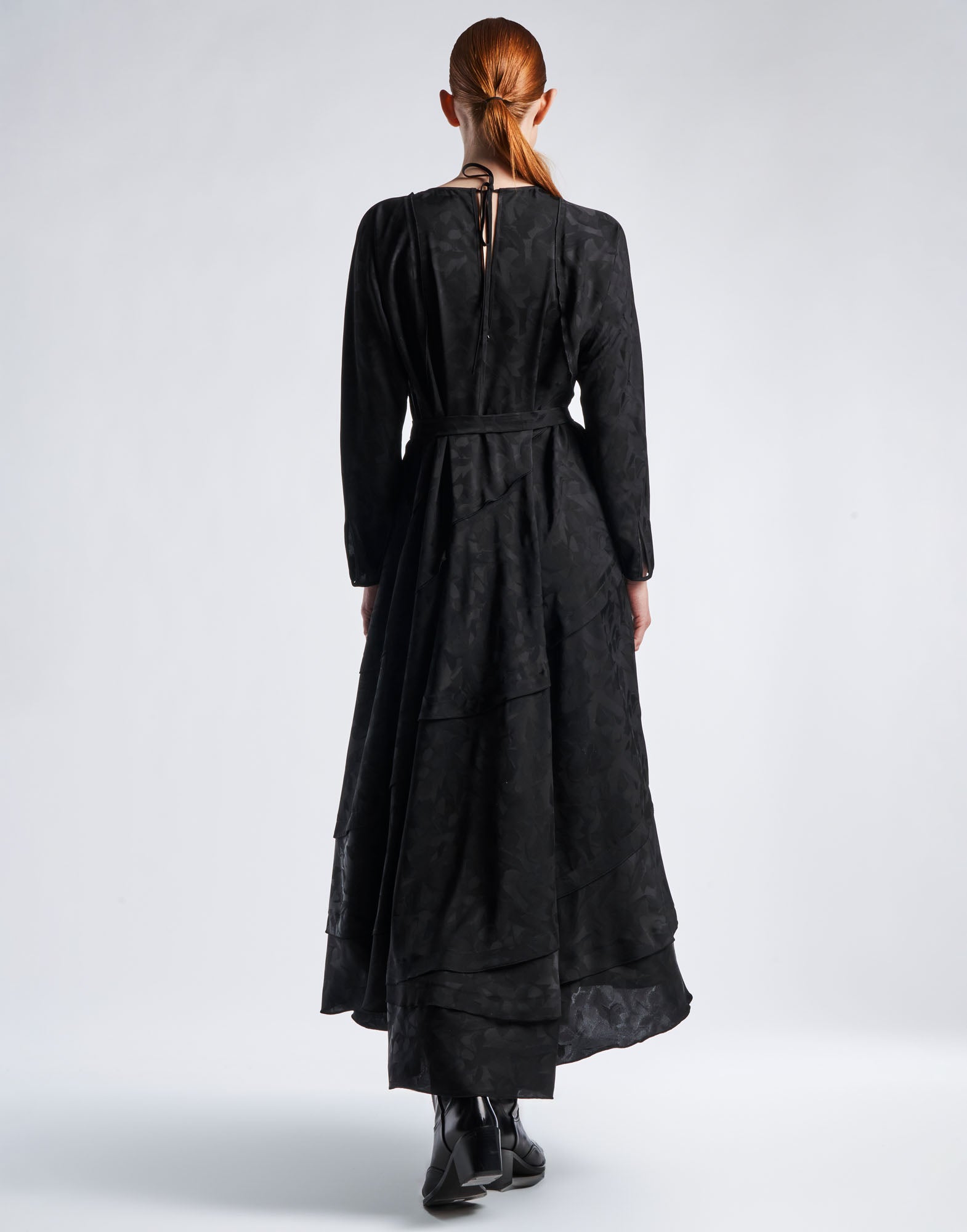 Rückansicht einer Person, die das schwarze Maxi-Kleid FAVOURITE aus Funktionssatin mit Damast-Blumenmuster, langen Dolmanärmeln und Schichten-Optik am Saum trägt.